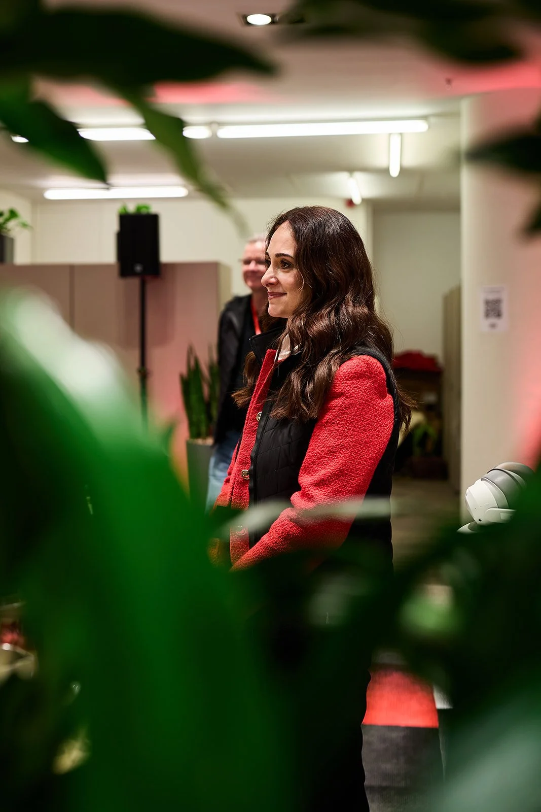 Eine lächelnde Frau mit braunen, gewellten Haaren steht in einem Büro, im Hintergrund eine Weitere Person, umgeben von Pflanzen und Büromöbeln.