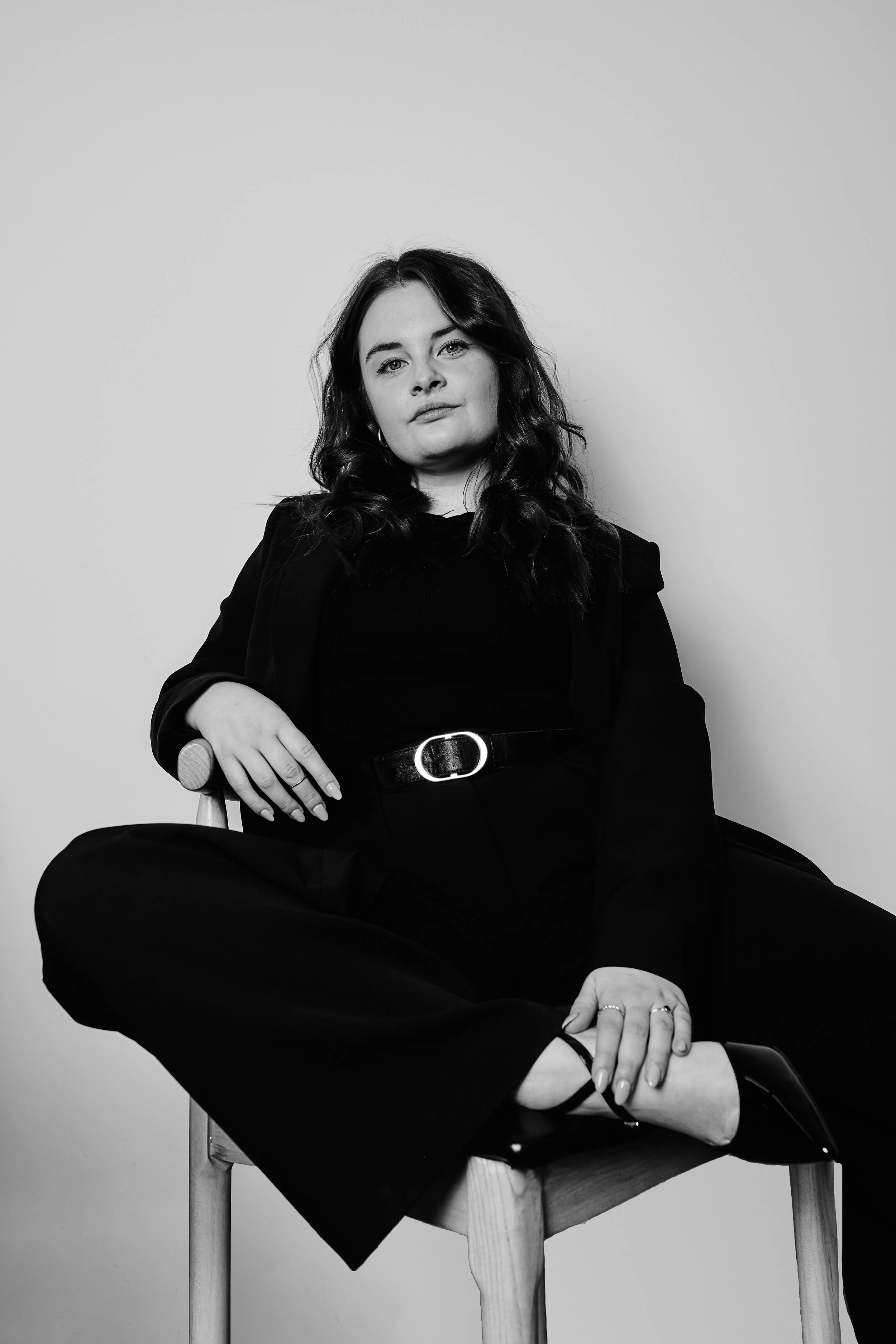 Schwarz-Weiß-Foto einer jungen Frau, die auf einem Stuhl sitzt, mit dunkler Kleidung und lockigen Haaren, vor einem einfachen hellen Hintergrund.