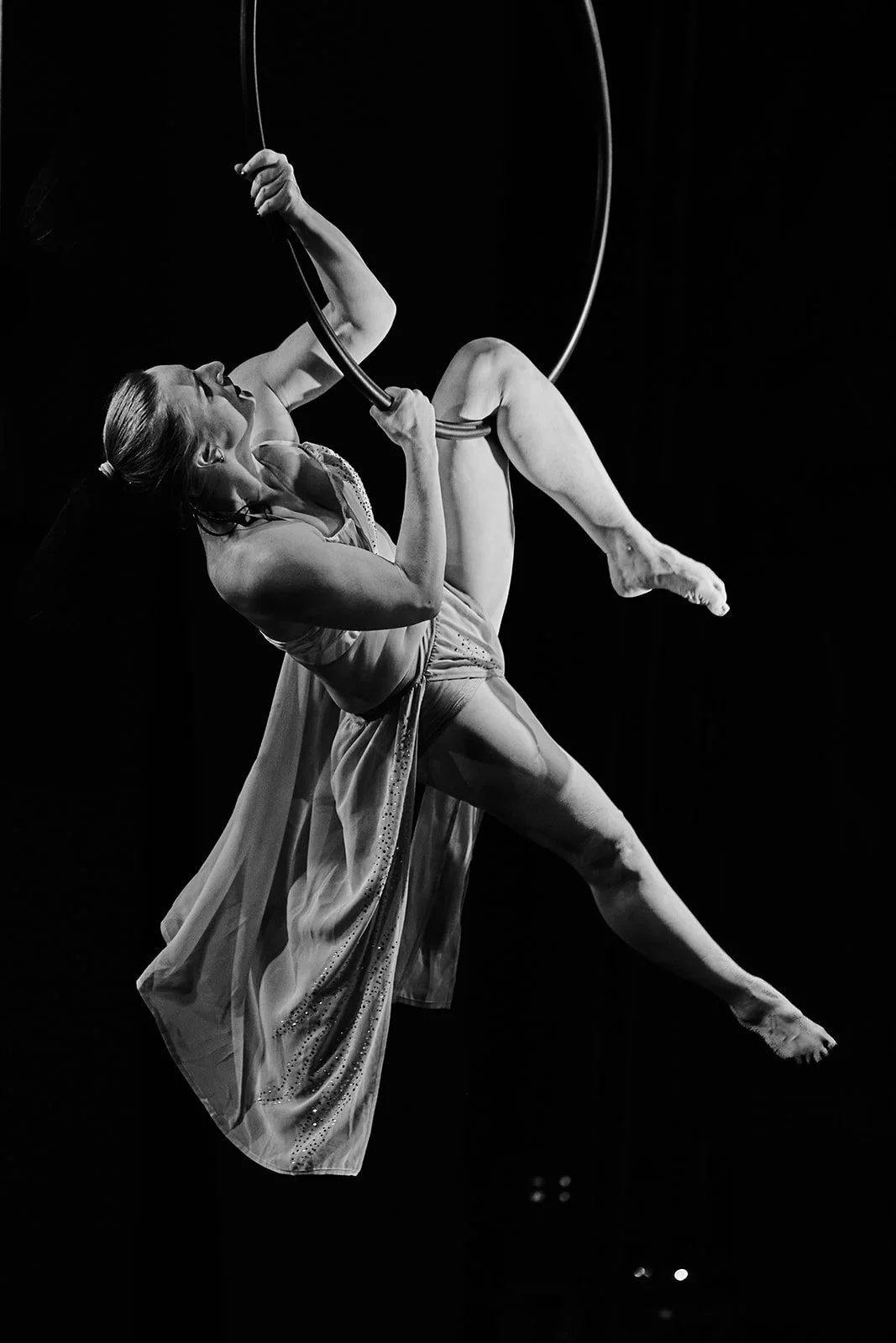 Poletänzerin im schwarzen Kleid schwingt an Ringen in der Luft, beide Beine um die Ringe gewickelt, in Schwarz-Weiß-Fotografie.