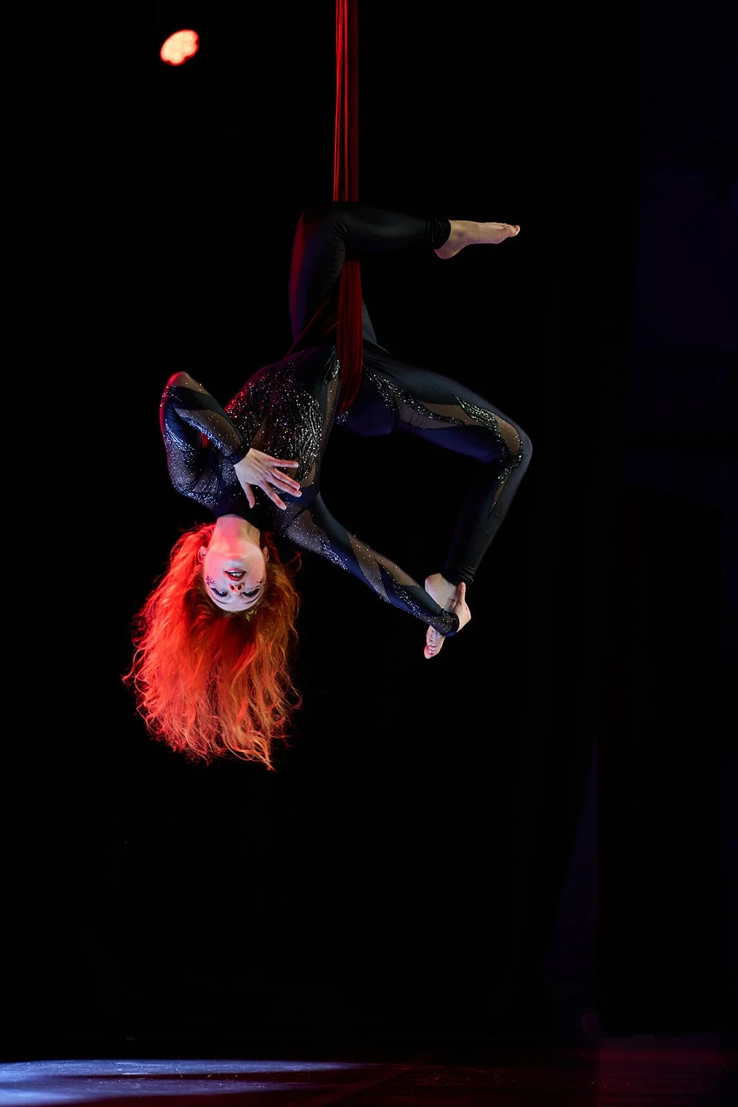 Akrobatin schwebt an einem roten Tuch in der Dunkelheit, trägt ein schwarzes, glitzerndes Kostüm mit transparenten Einsätzen und leuchtend rotes, lockiges Haar.