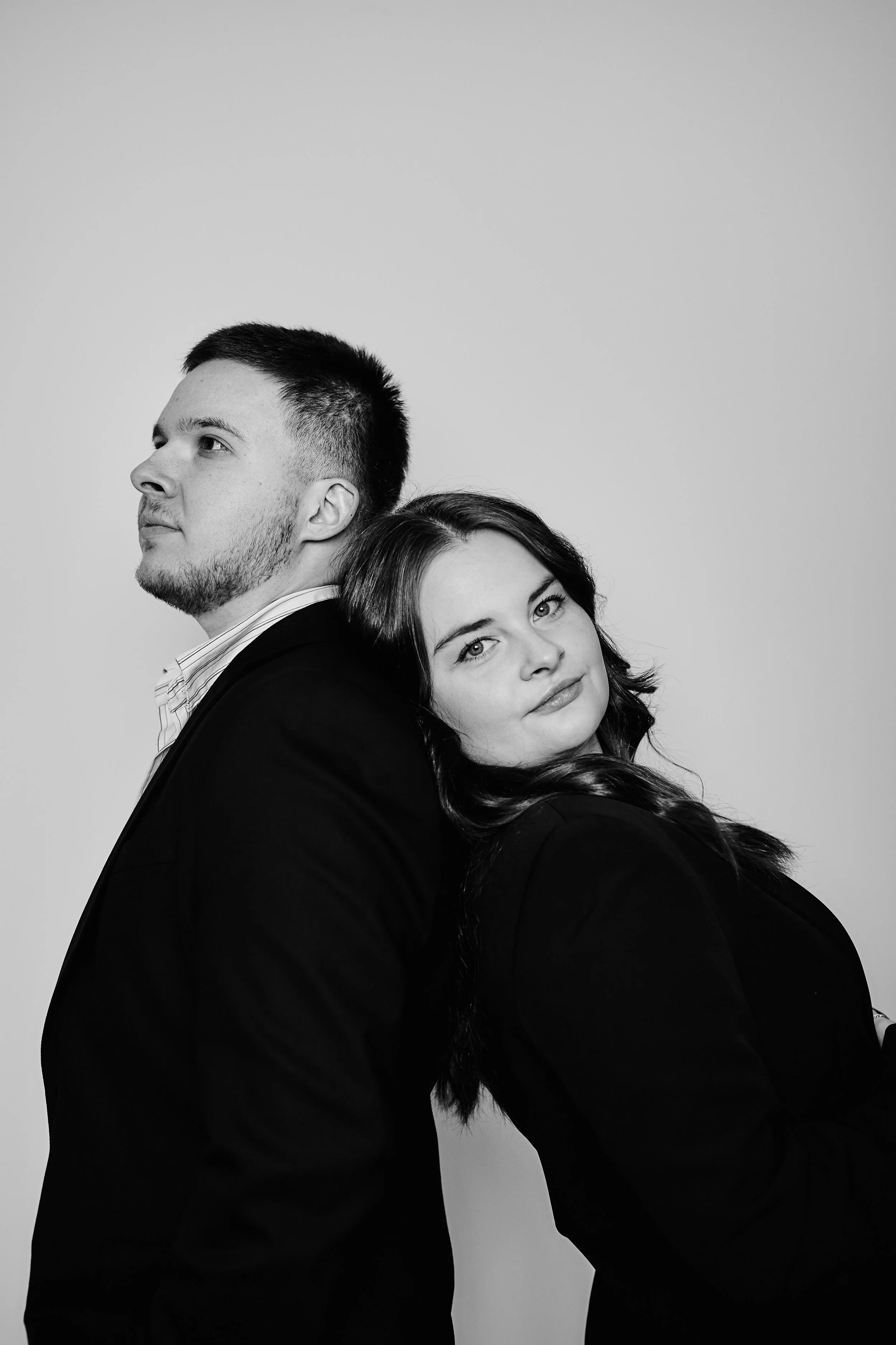 Schwarz-weiß-Fotografie eines Mannes und einer Frau, die Rücken an Rücken stehen, mit entspanntem Blick in die Kamera, lässige Kleidung auf hellem Hintergrund.