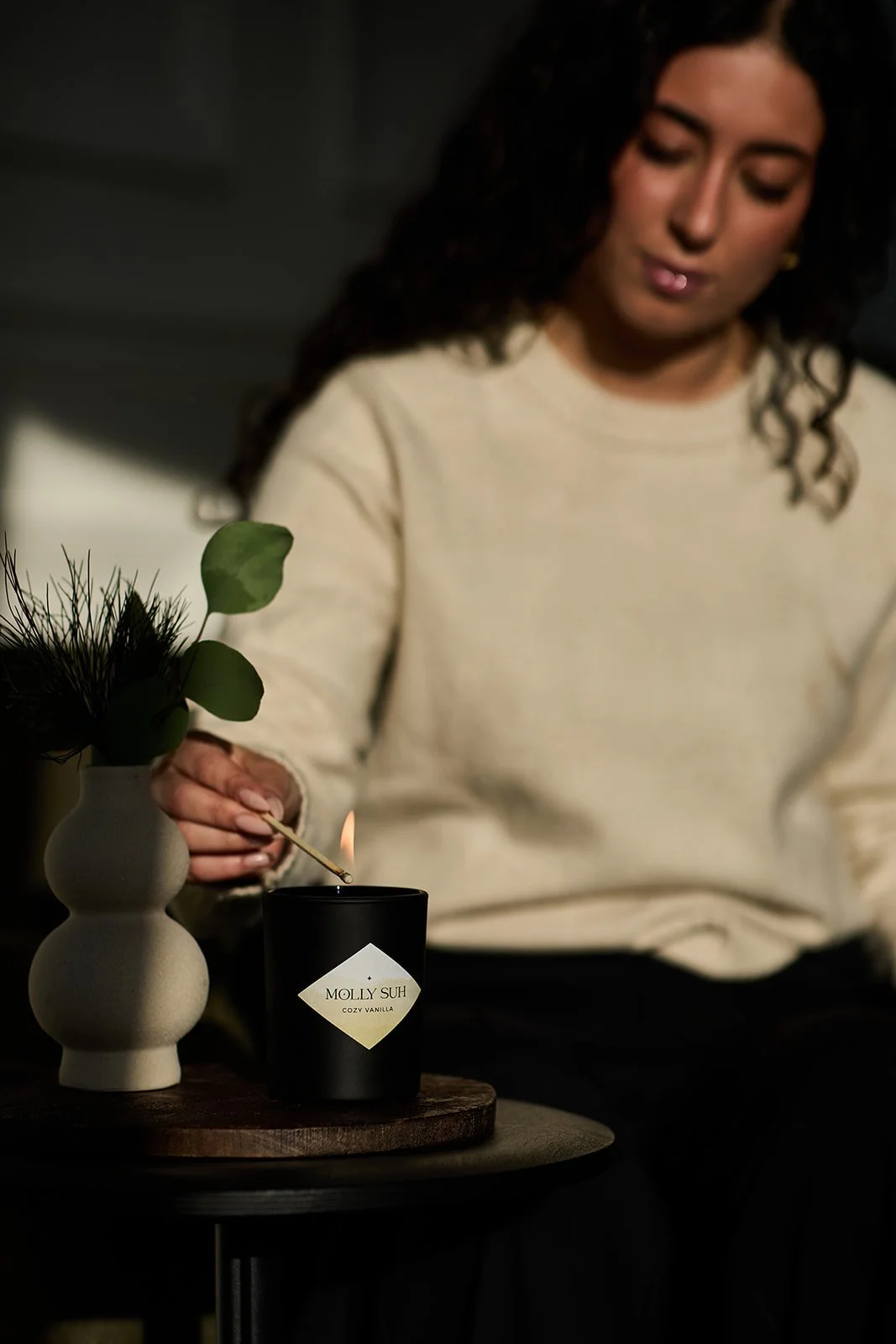 Eine Frau zündet eine Kerze in einem schwarzen Behälter an, der mit dem Etikett 'MOLLY SUH COZY VANILLA' versehen ist, auf einem dunklen Tisch neben einer weißen Vase mit Blättern.