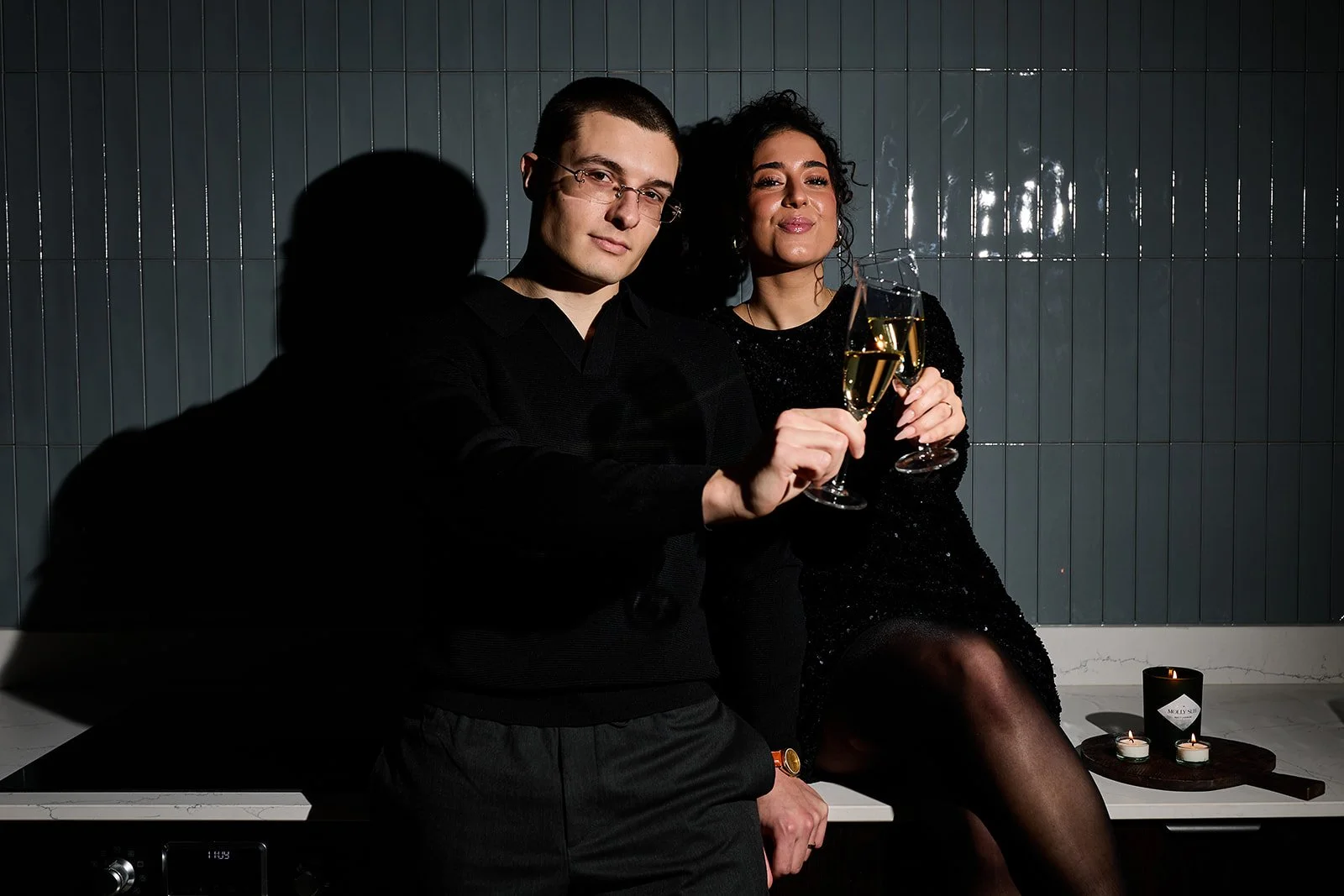 Ein Mann und eine Frau feiern mit Sektgläsern in einer modernen, dunklen Umgebung, Sektempfang, Kerzen und schwarze Kleidung