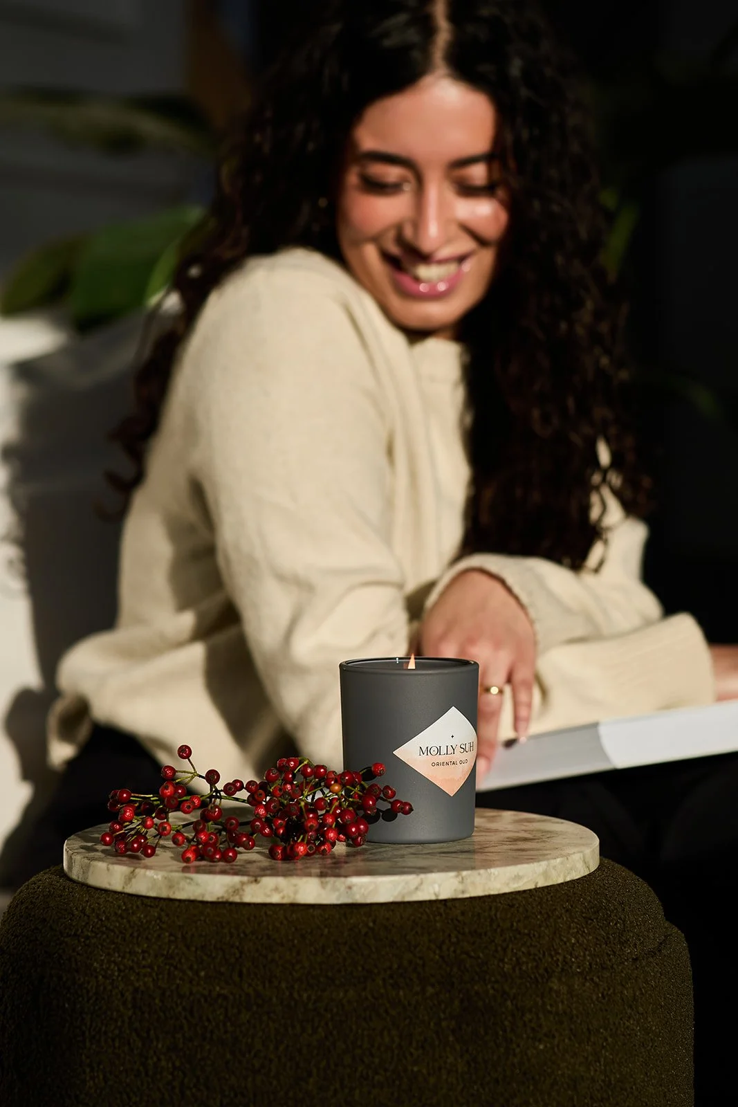 Eine lachende Frau mit lockigem Haar trägt eine cremefarbene Strickjacke und sitzt hinter einem kleinen runden Tisch. Auf dem Tisch steht eine schwarze Kerze mit einem Etikett, auf dem "MOLLY SUG" steht, neben einer Zweig mit roten Beeren.