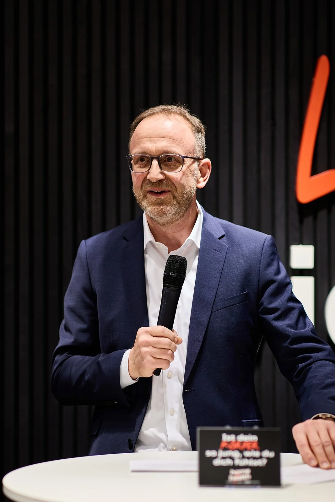 Ein Mann mit Brille trägt einen dunklen Anzug und hält ein Mikrofon in der Hand, während er an einem Tisch sitzt. Im Hintergrund befindet sich eine dunkle Wand mit einer großen roten Zahl oder Buchstabe.