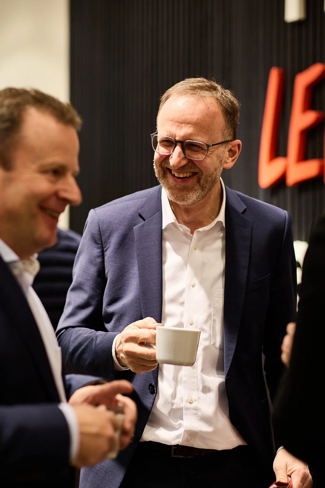 Zwei Männer im Gespräch bei einer Veranstaltung. Einer hält eine Tasse, beide lachen.