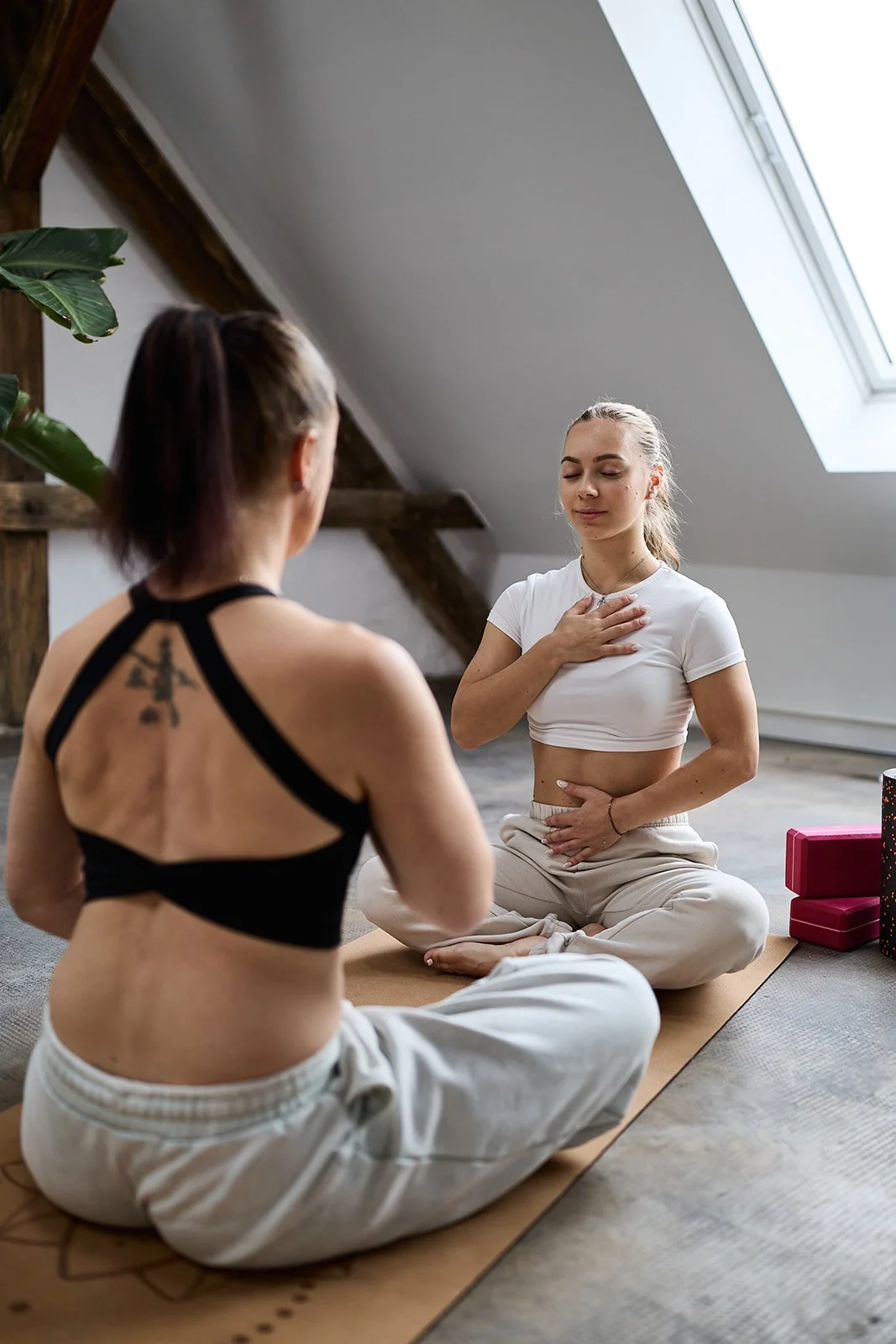 Zwei Frauen praktizieren Yoga in einem hellen Raum unter einem Dachfenster, sitzend im Schneidersitz auf einer Yogamatte.