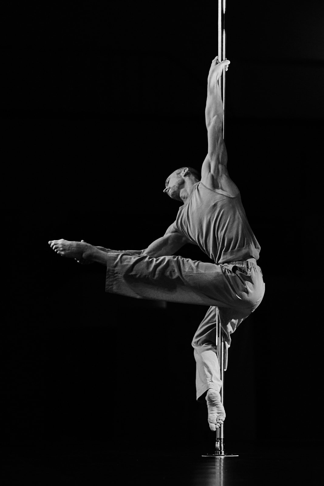 Ein männlicher Akrobat führt eine Pole-Akrobatik-Nummer auf, hängt an einem vertikalen Stab, in einem schwarzen Raum, schwarz-weißes Foto.