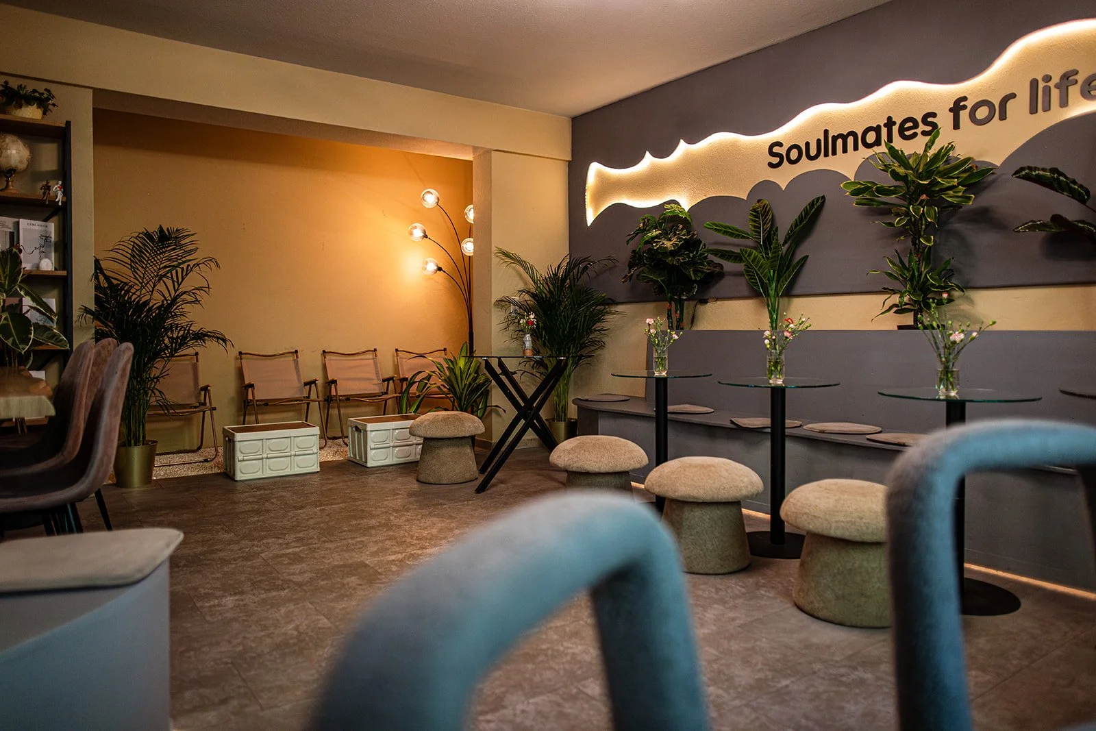 Ein modern eingerichteter Raum mit Stühlen und Tischen, Dekorpflanzen und einer Regalwand. An der Wand ist ein Leuchtschild mit der Aufschrift 'Soulmates for life'.