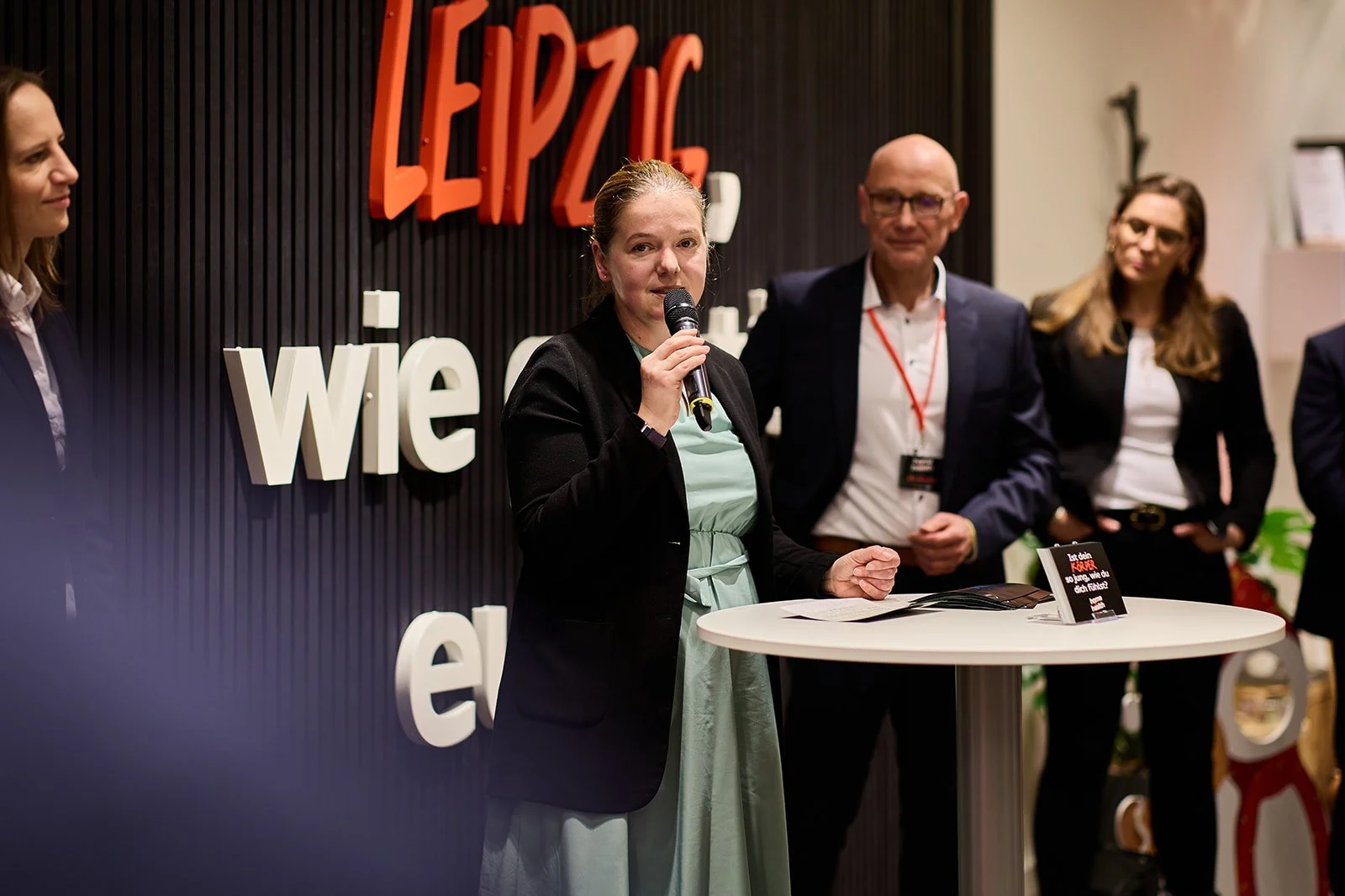 Eine Gruppe von Menschen bei einer Veranstaltung, bei der eine Frau mit einem Mikrofon spricht, im Hintergrund ist ein Wand mit großen Buchstaben und der Schriftzug "LEIPZIG" sichtbar.