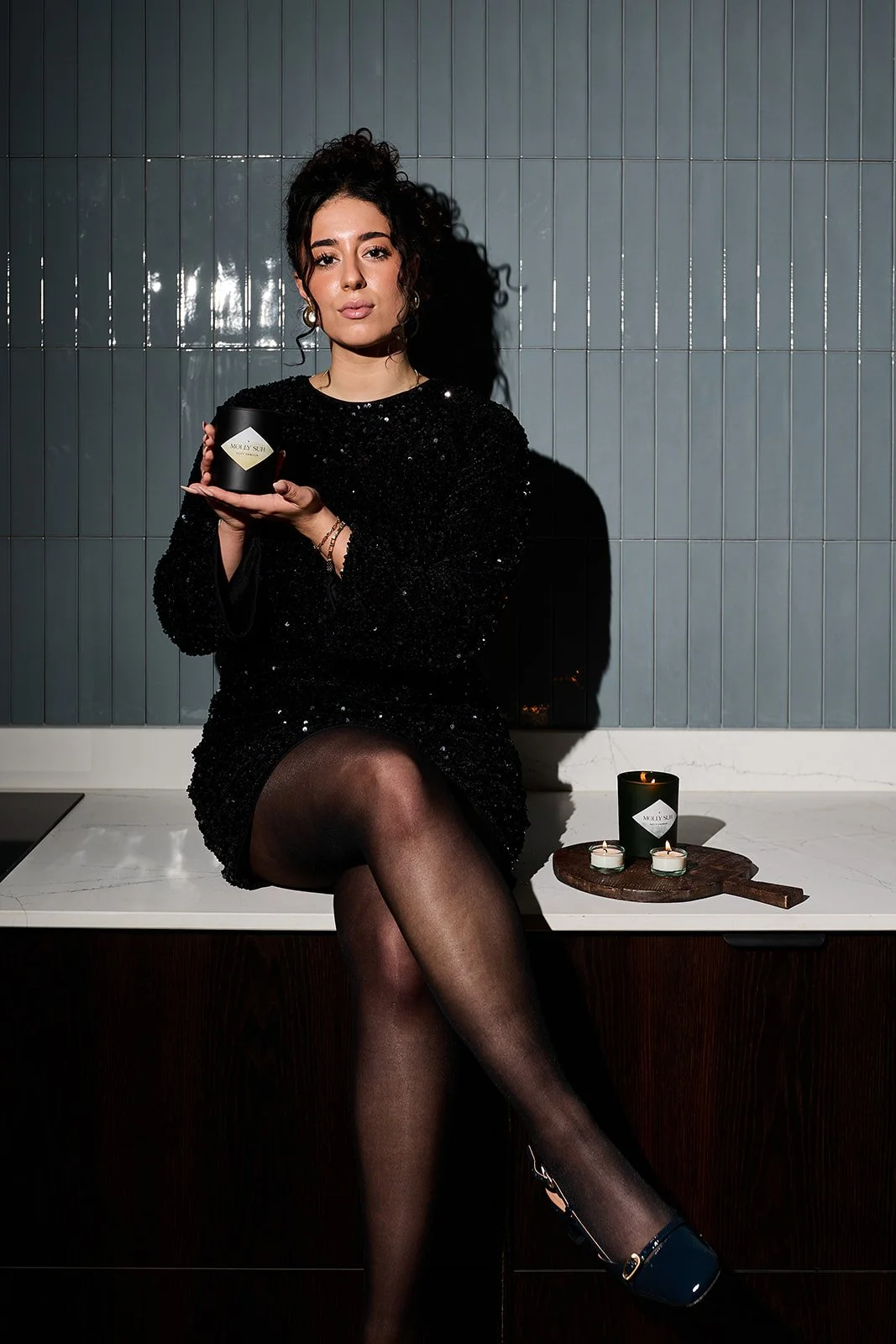 Eine Frau in einem schwarzen, glänzenden Kleid sitzt auf einer Küchenarbeitsplatte und hält eine schwarze Kerze in den Händen. Es gibt zwei weitere Kerzen auf einem Holzbrettchen nebeneinander.