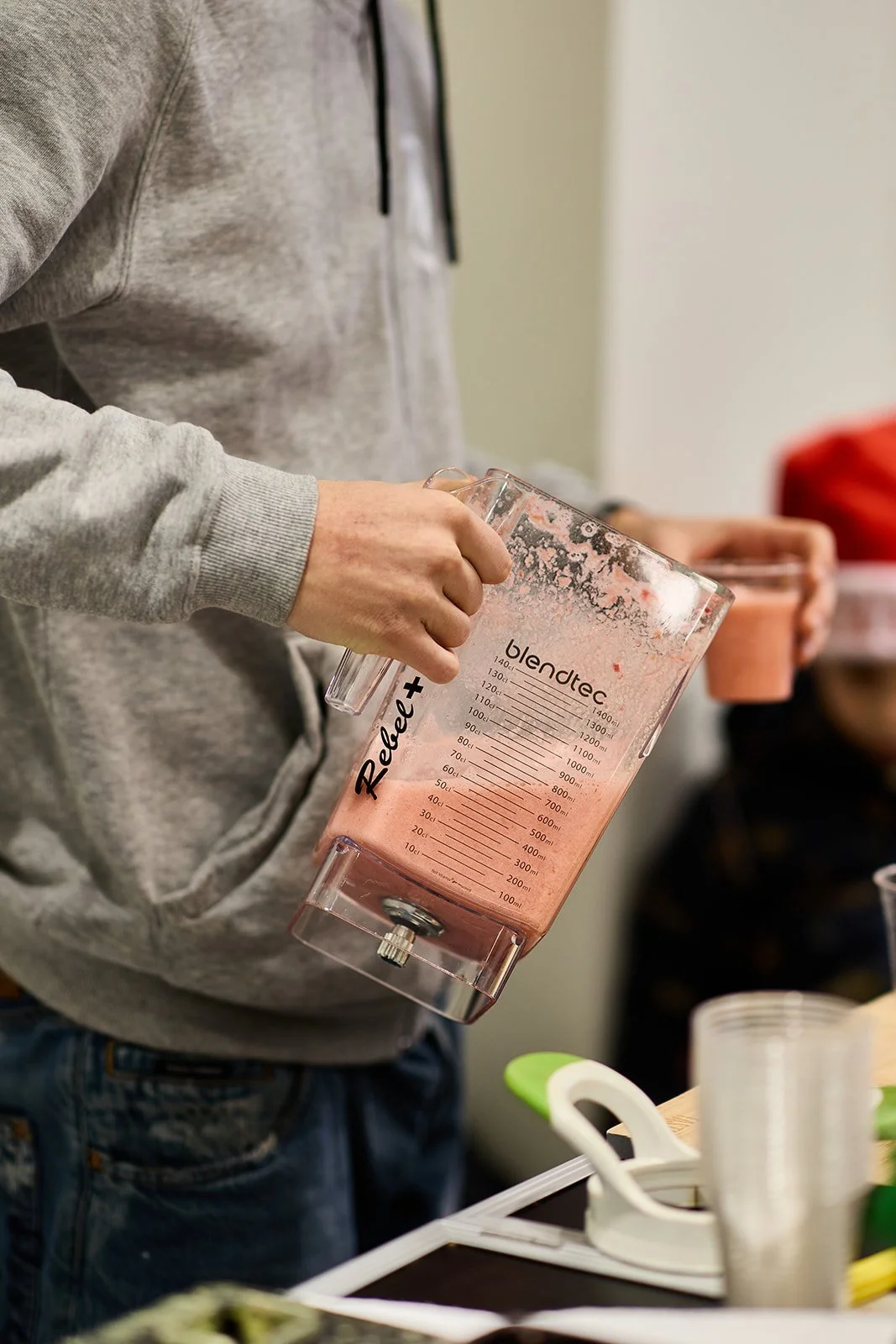 Person hält einen großen, transparenten Mixer mit pinkem Getränk. Im Hintergrund andere Personen, die ebenfalls Getränke halten, und Küchenutensilien auf dem Tisch.