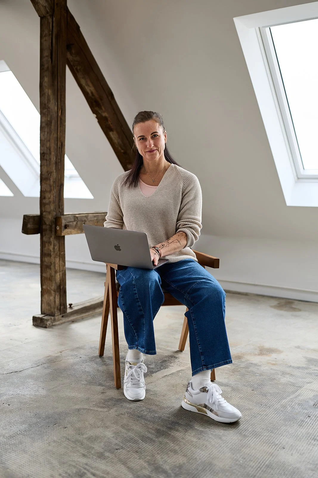 Eine junge Frau sitzt auf einem Stuhl vor einem weißen Wand im Dachgeschoss mit großen Skylight-Fenstern, arbeitet an einem Laptop.