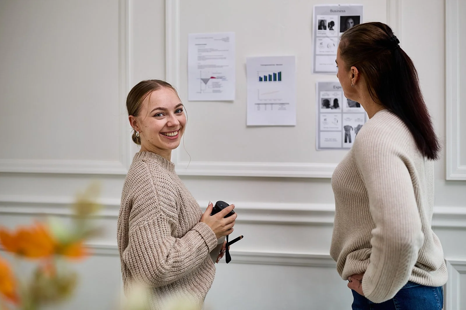 Zwei Frauen sprechen vor einer Pinnwand mit Dokumenten und Grafiken in einem Büroraum.