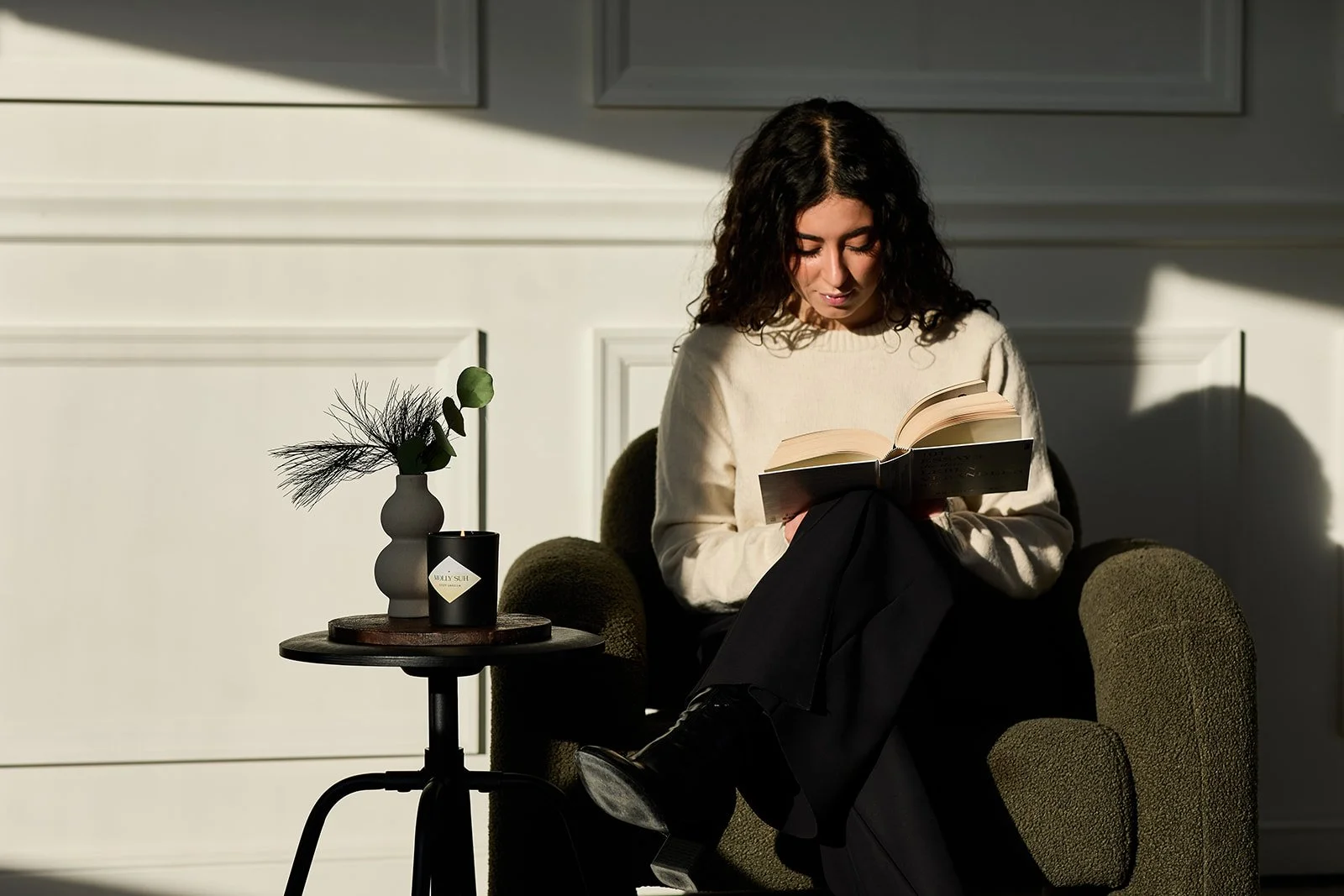 Junge Frau sitzt in einem Sessel, liest ein Buch, neben ihr ein kleiner Tisch mit einer Vase und einer Kerze, im Hintergrund weiße Wand mit Schatten