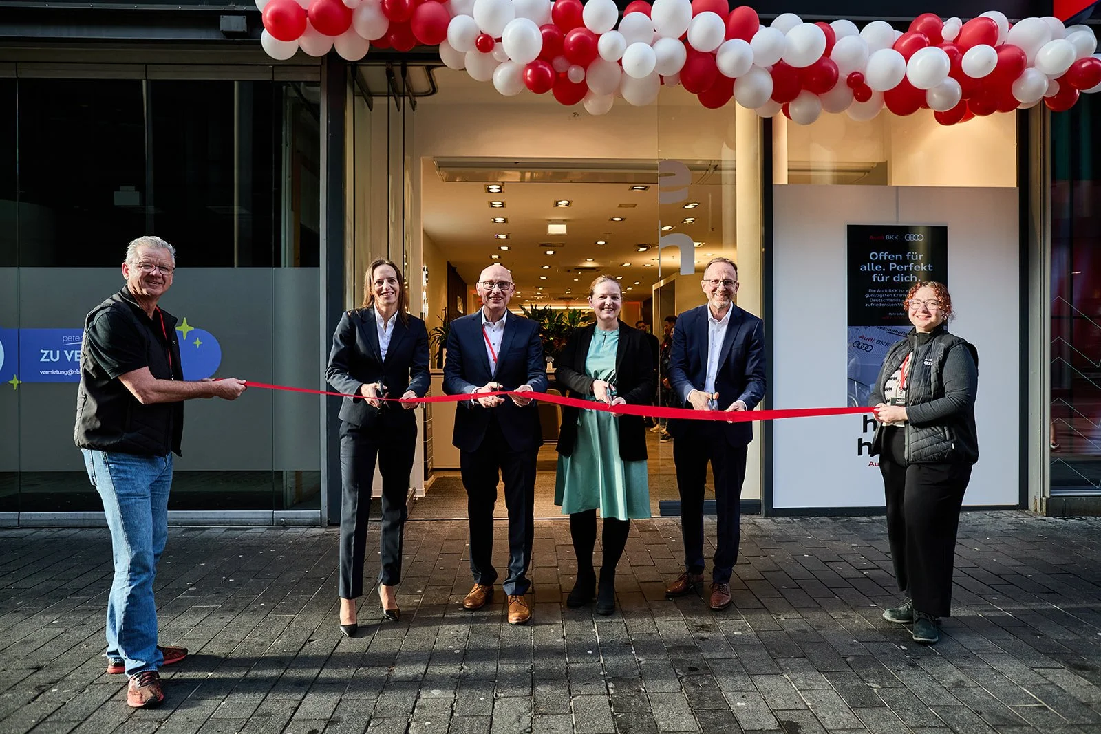 Eine Gruppe von sechs Menschen beim Eröffnungsrieß im Eingangsbereich eines Gebäudes, mit roten und weißen Ballons an der Decke. Sie halten zusammen eine rote Schleife, während sie nach links und rechts blicken und lächeln.