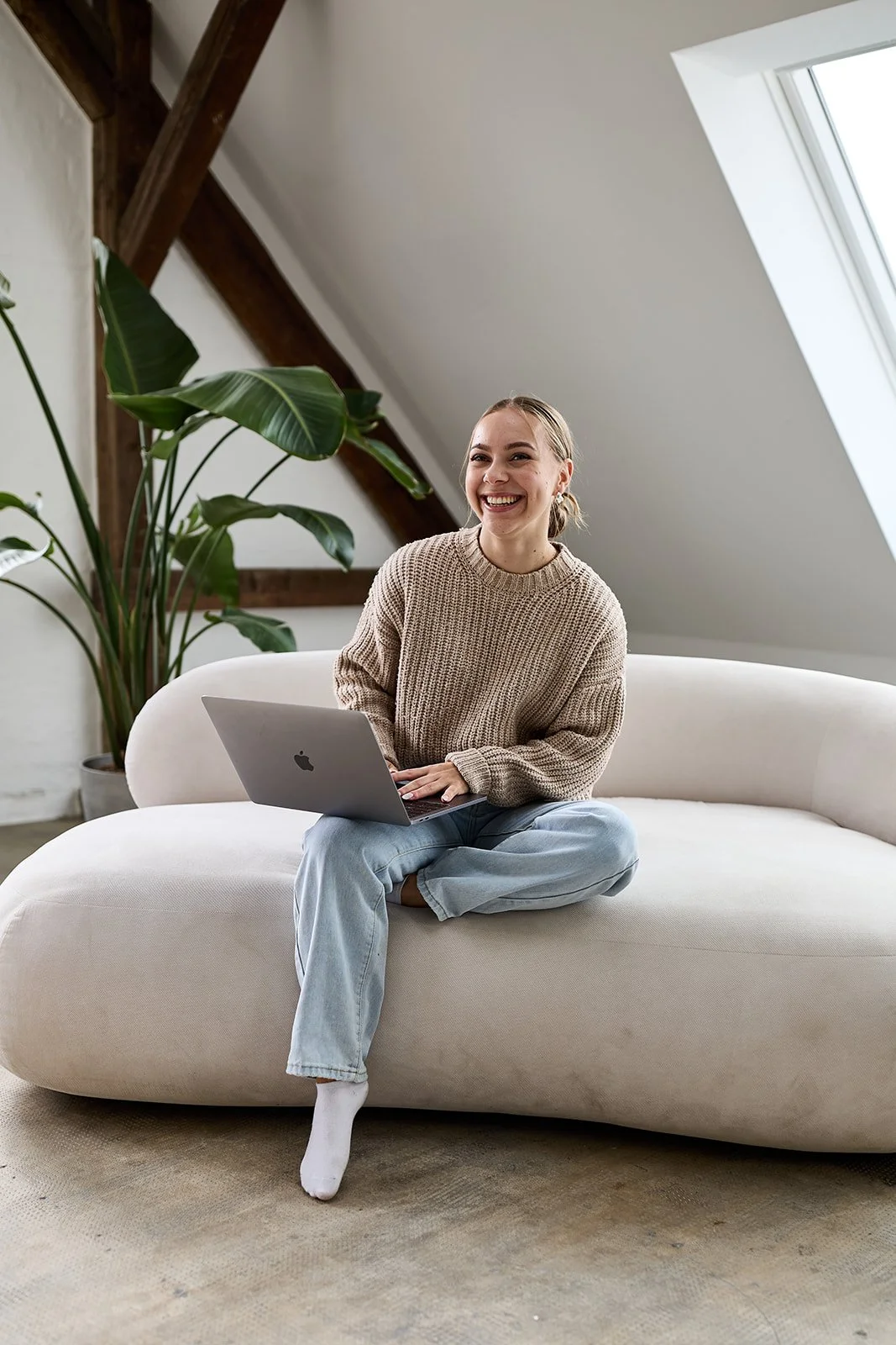 Junge Frau sitzt auf einem weißen Sofa mit einem Laptop in einem hellen Raum mit großem Fenster und Pflanzen.