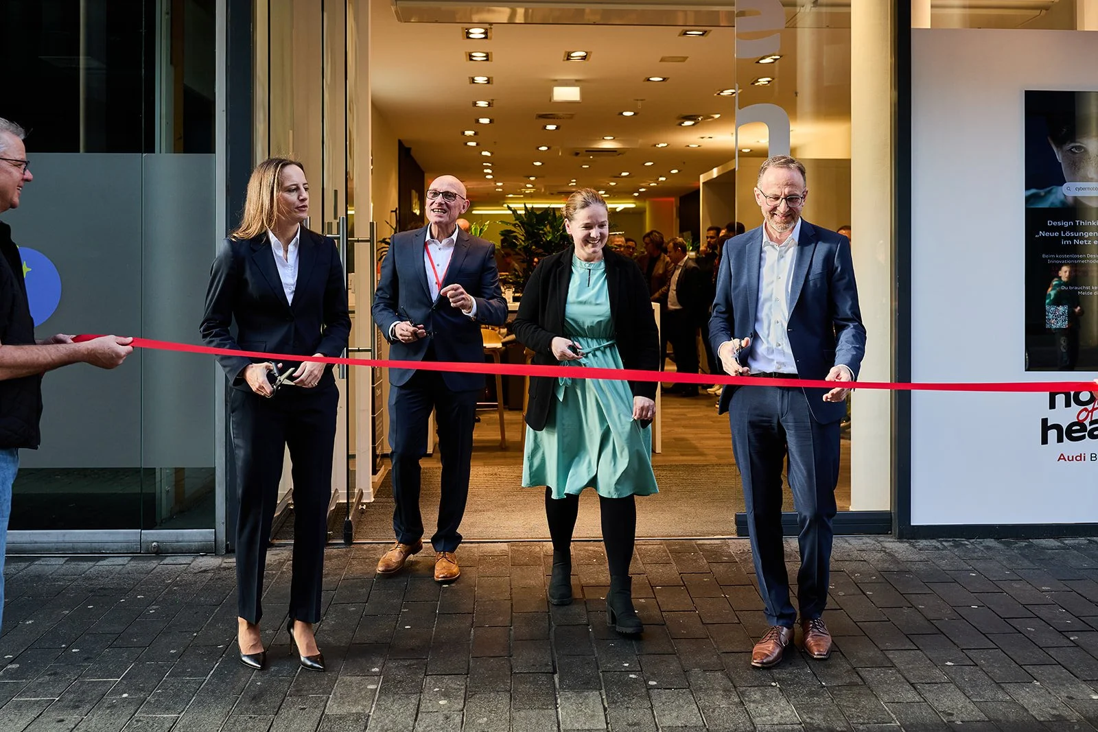 Eine Gruppe von fünf Menschen beim Eröffnungszeremonie, die einen roten Band durchschneiden. Sie stehen vor einem Eingang zu einem Gebäude, vermutlich ein Büro oder Veranstaltungsraum, bei Tag oder Abend.