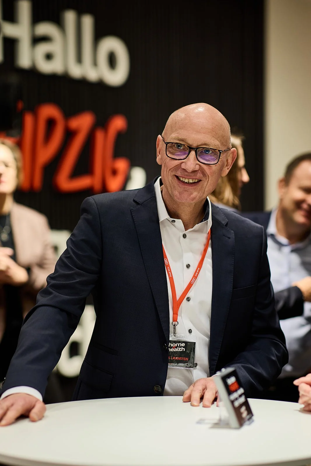 Mann im Geschäftsanzug mit Brille und Namensschild, lächelnd und an einem weißen Tisch stehend, hinter ihm eine Gruppe von Menschen in einem Besprechungsraum mit einer Schwarzwand, die die Wörter 'hallo' und 'PIZZA' zeigt.