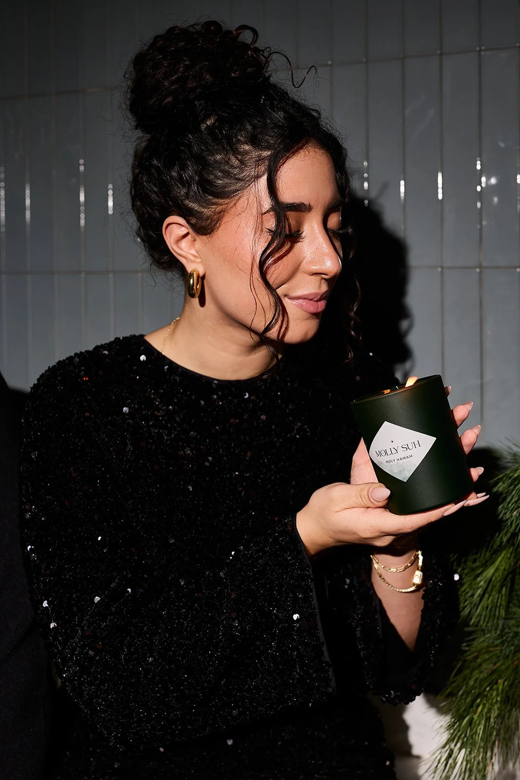 Eine Frau mit dunklen, gelockten Haaren in einem schwarzen, glitzernden Kleid hält eine schwarze Kerze mit einem weißen Etikett, auf dem 'MOLLY SUH' geschrieben steht. Die Frau trägt goldene Ohrringe und Armbänder.