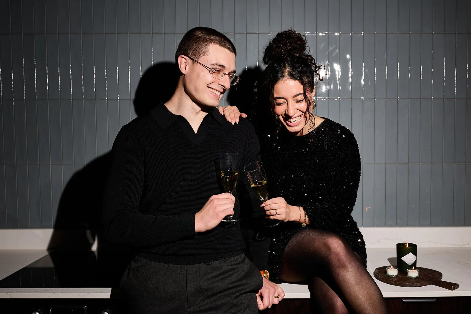 Ein Mann und eine Frau sitzen auf einer Küchenarbeitsplatte, lachen und stoßen mit Sektgläsern an. Sie tragen dunkle Kleidung, und im Hintergrund sind Kerzen auf einem Schneidebrett zu sehen.