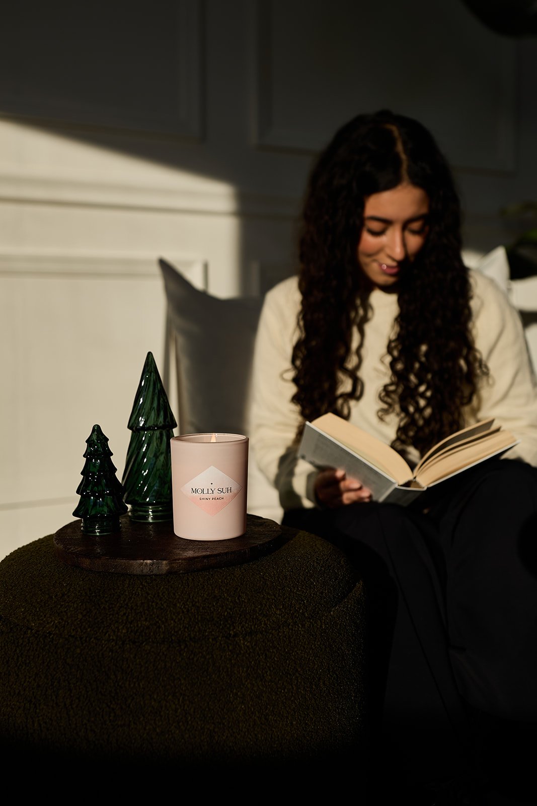 Junge Frau liest ein Buch, sitzend neben einer dekorativen Weihnachtsbaum- und Kerzendekoration, mit Schatten und Licht auf der Wand im Hintergrund.