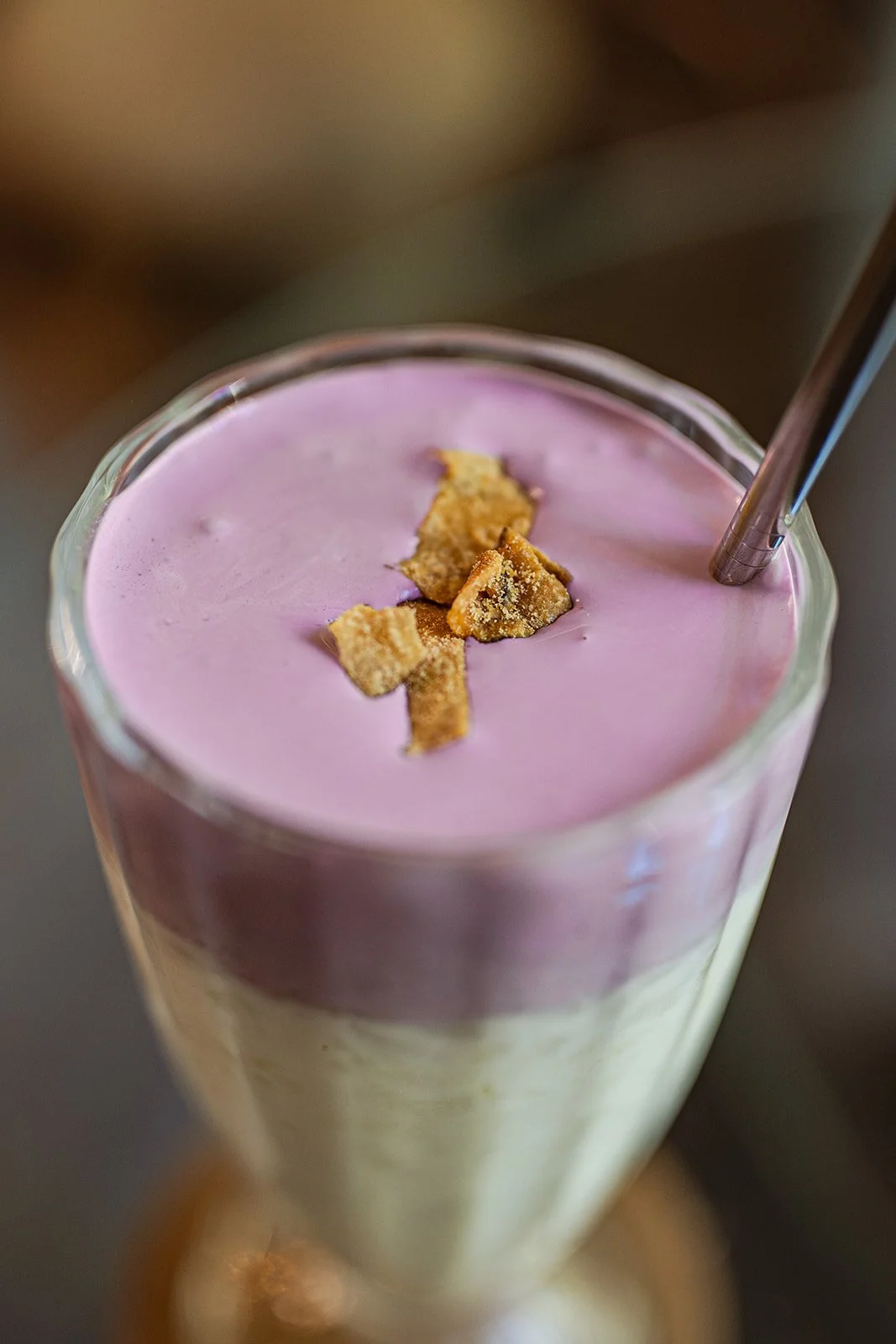 Ein Glas mit einer zweifarbigen Schicht, oben rosa und unten hellgelb, mit knusprigen Kekskrümeln und einem Strohhalm.