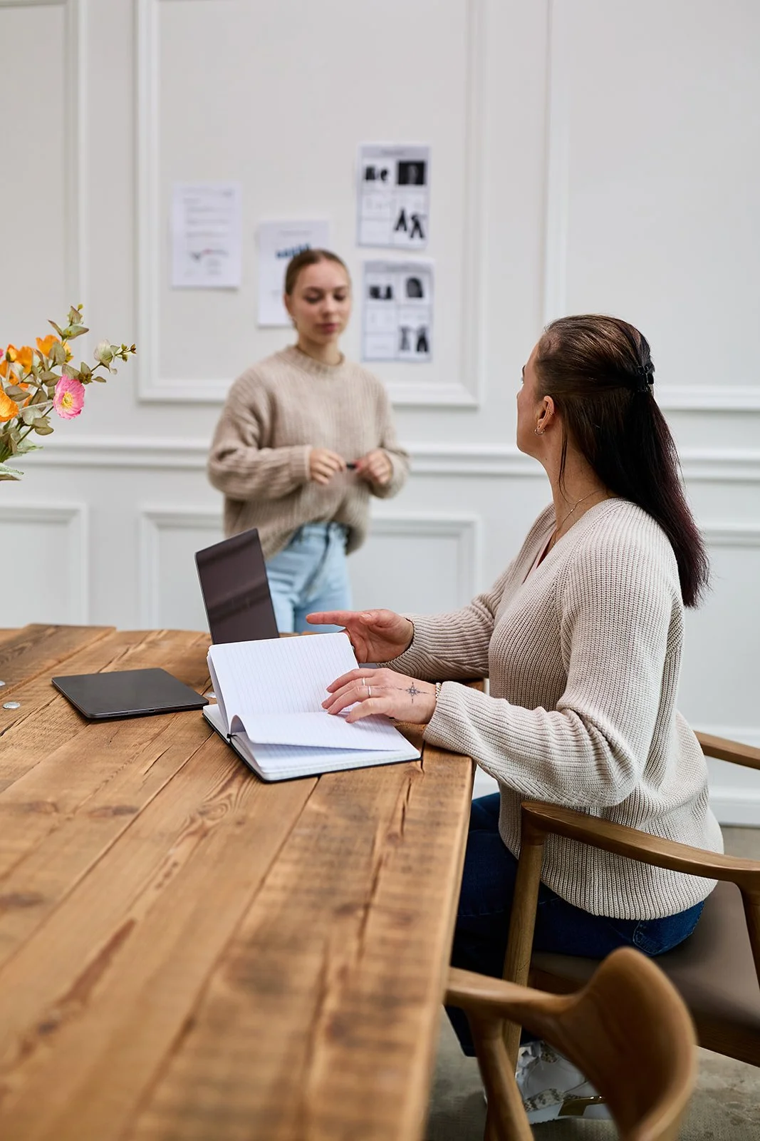 Zwei Frauen in einem Büro, die an einem Holztisch sitzen und sprechen. Eine Frau macht Notizen in einem Notizbuch, während die andere steht.