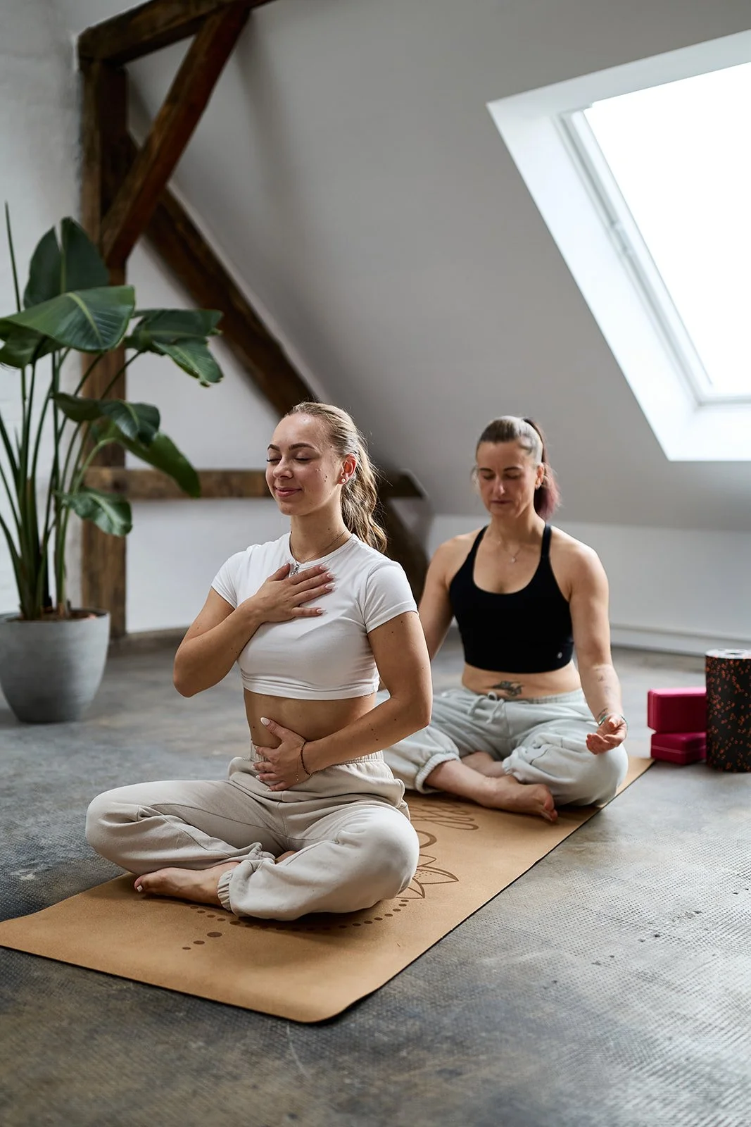 Zwei Frauen beim Yoga- oder Meditationspraktikum in einem hellen Raum mit Dachfenster, eine sitzt mit geschlossenen Augen und Hand auf der Brust, die andere hinter ihr, in einer Meditationshaltung.
