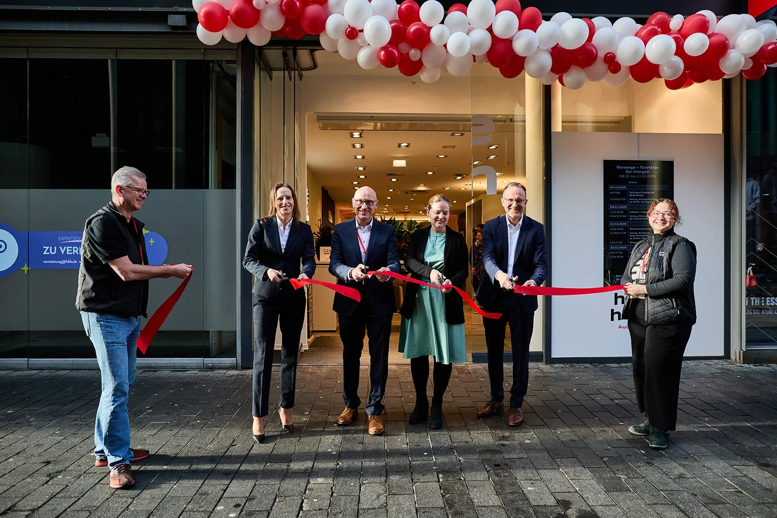 Menschen bei einer Eröffnungskundgebung vor einem Geschäft mit roten und weißen Luftballons, die eine rote Schleife durchschneiden.