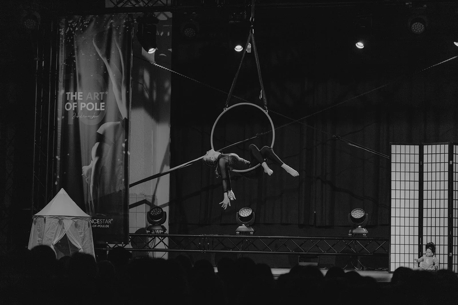 A Zirkus-Performance mit Akrobatin, die an einem Ring in der Luft hängt, auf einer Bühne mit dunklem Hintergrund. Es gibt Bühnenlichter, ein Zelt und ein Kind im Hintergrund.