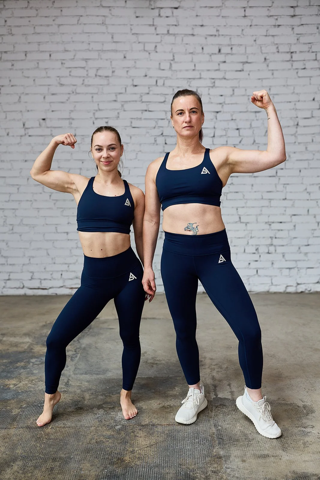 Zwei Frauen in Sportkleidung, die stolz ihre Muskeln zeigen, stehen vor einer weißen Ziegelwand.