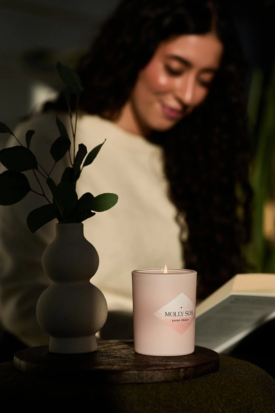 Eine Kerze mit dem Label 'MOLLY SUH SHINY PEACH' brennt, daneben eine Vase mit grünen Zweigen, im Hintergrund eine lachende Frau mit dunklen, lockigen Haaren, die ein Buch liest.