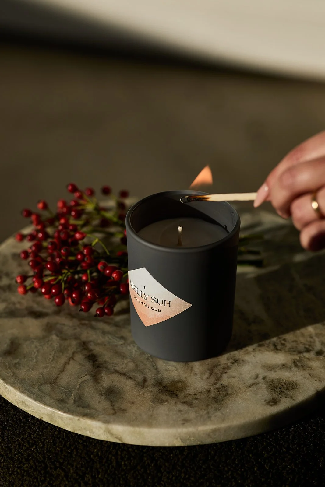 Person zündet eine schwarze Kerze mit einem Matchstick an, auf einem grauen Stein-Tablett mit Roten Beeren als Dekoration.
