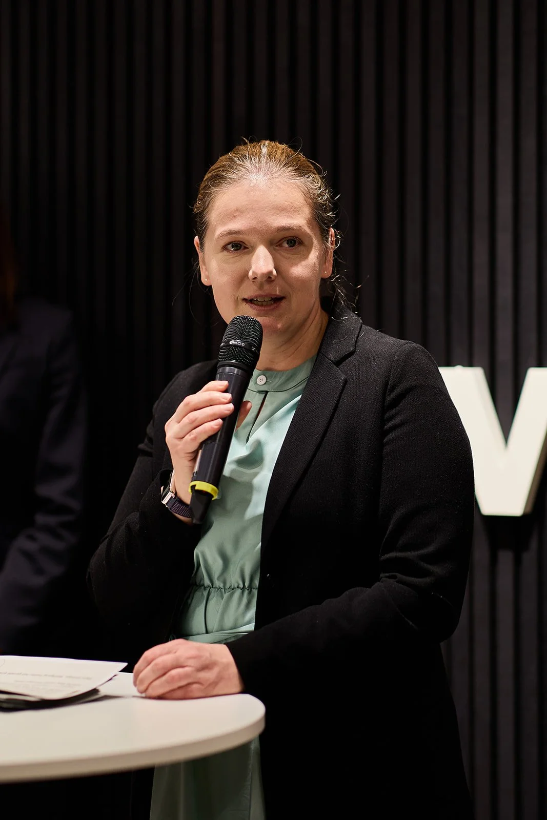 Eine Frau mit braunem Haar, die eine schwarze Jacke und ein grünes Kleid trägt, hält ein Mikrofon in der Hand und spricht vor einem schwarzen Hintergrund mit weißen Buchstaben.