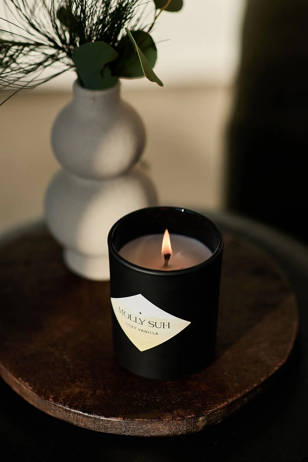 Schwarze Kerze mit Flamme, beschriftet mit 'MOLLY SUH COZY VANILLA', auf einem runden Holztablett, im Hintergrund eine weiße Vase mit grünen Blättern und Zweigen.