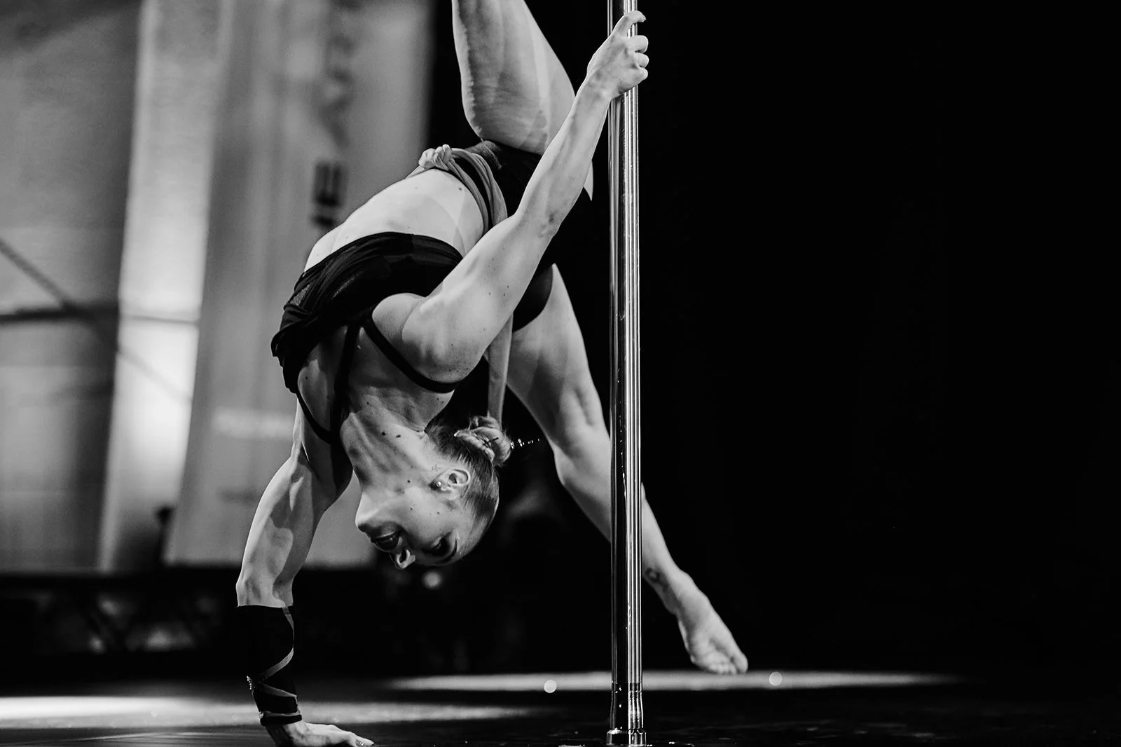Eine Frau führt eine Pole Dance-Performance auf einer Bühne aus, sichtbar von der Seite. Sie befindet sich in einer aufwändigen Pose, bei der sie sich an einer vertikalen Stange festhält und auf dem Boden balanciert.