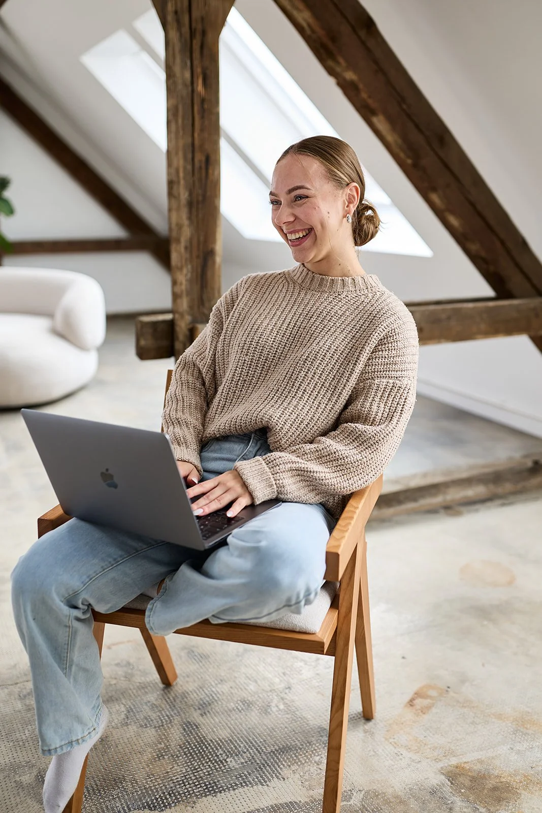 Eine lachende Frau sitzt auf einem Stuhl mit einem Laptop auf ihrem Schoß in einem modernen, hellen Raum mit Holzbalken und großen Fenstern im Dach.
