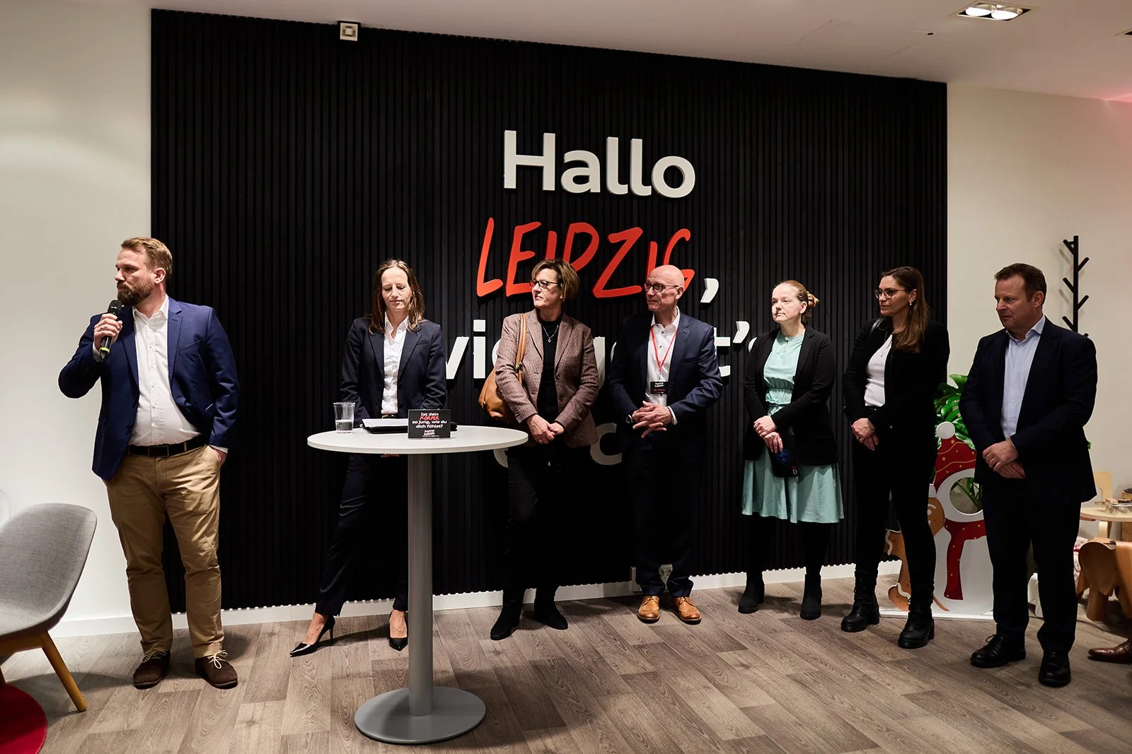 Sieben Menschen stehen vor einer schwarzen Wand mit der Aufschrift "Hallo LEIPZIG, willkommen". Ein Mann auf der linken Seite hält ein Mikrofon, die Menschen sind in formelle Kleidung. Es gibt einen Stehtisch und einen Weihnachtsbaum-Dekor im Raum.