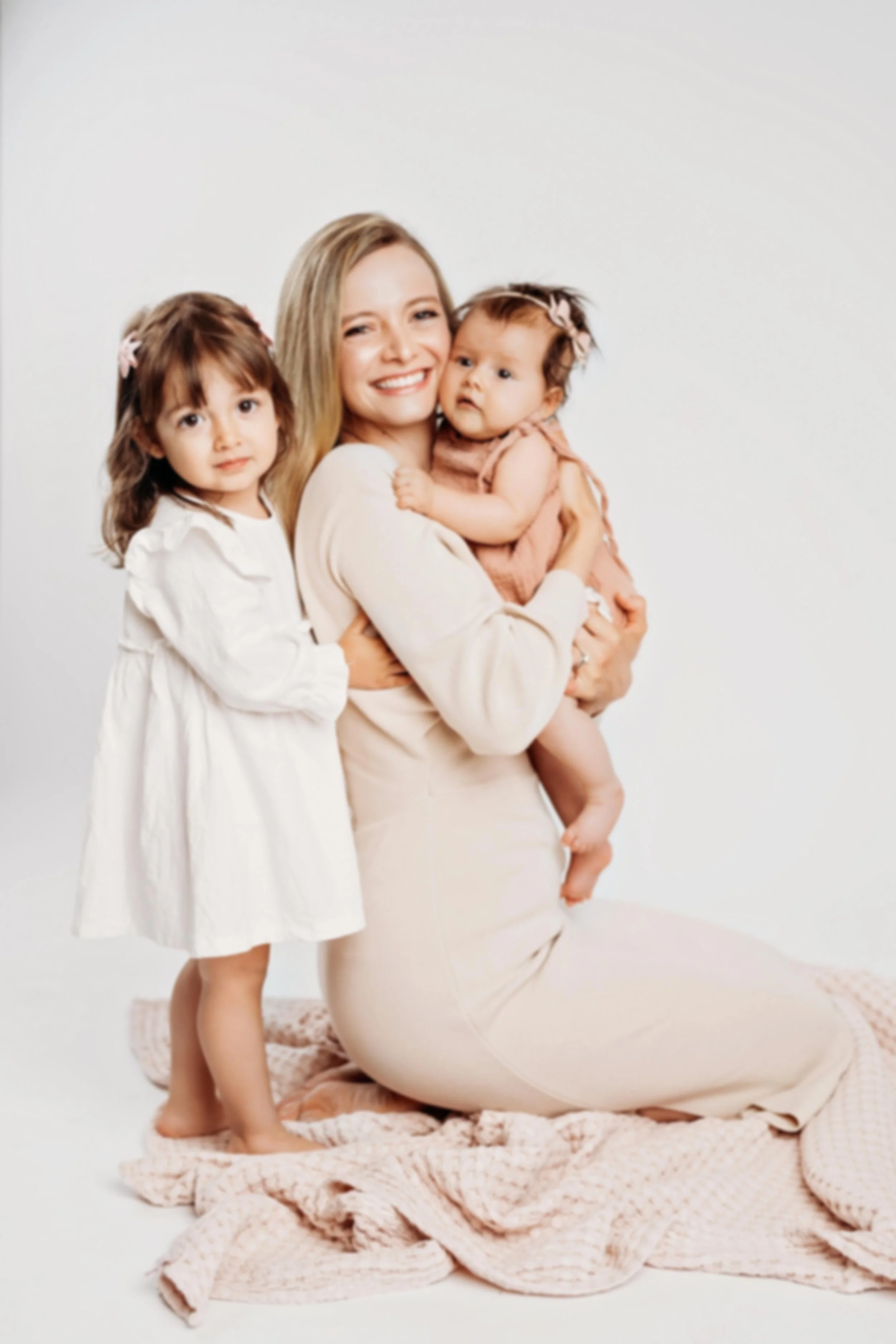 A woman, mom, with two young girls, one standing and one being held, all smiling against a plain white background. loving, caring. family bond.