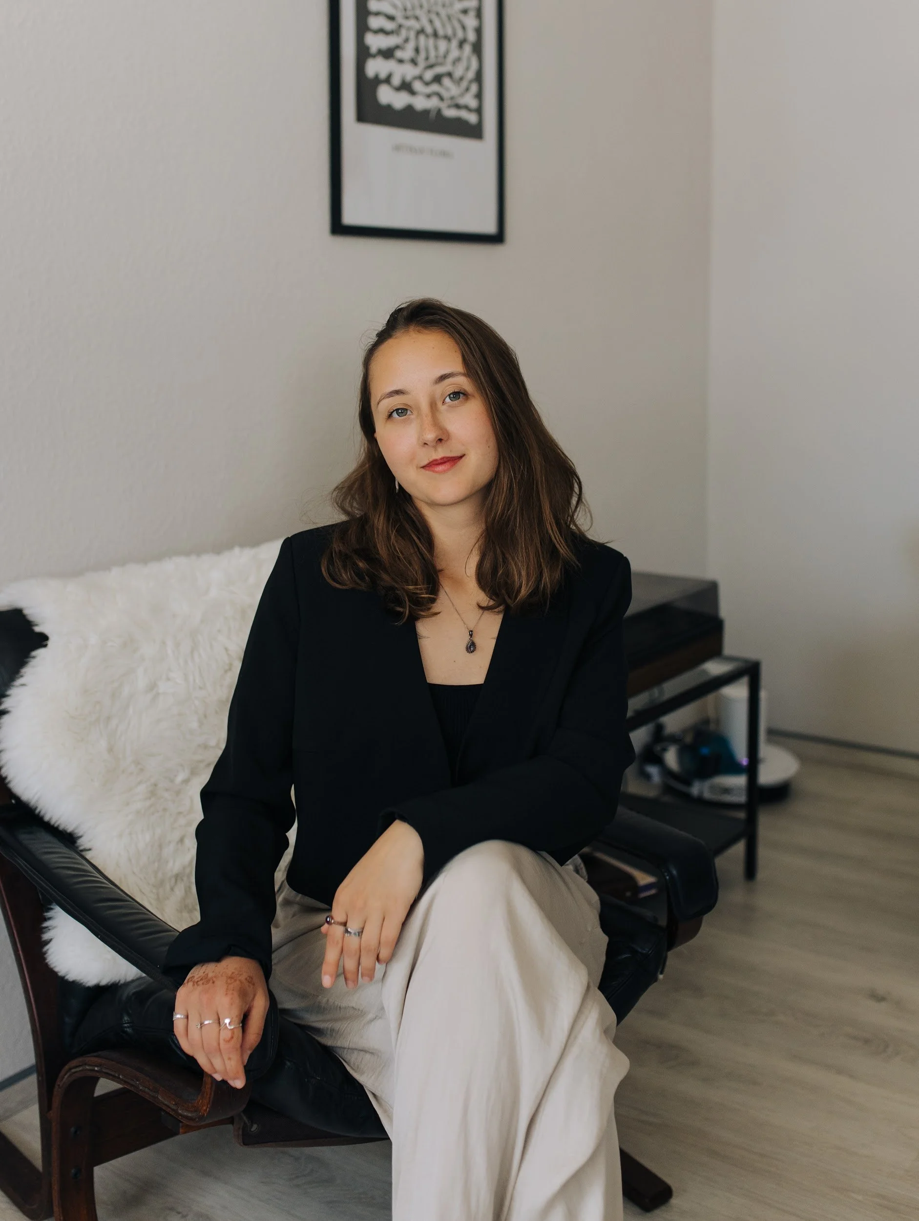 Junge Frau mit braunen Haaren, schwarzer Jacke, cremfarbener weiten Hose und Schmuck sitzt auf einem Stuhl in einem Raum, im Hintergrund ein Bild an der Wand und ein tabletop mit elektronischen Geräten.