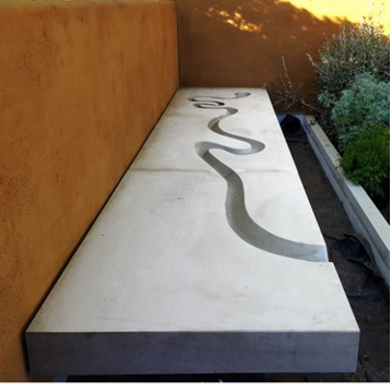 Stone bench with a curved groove, next to an orange wall with plants nearby.