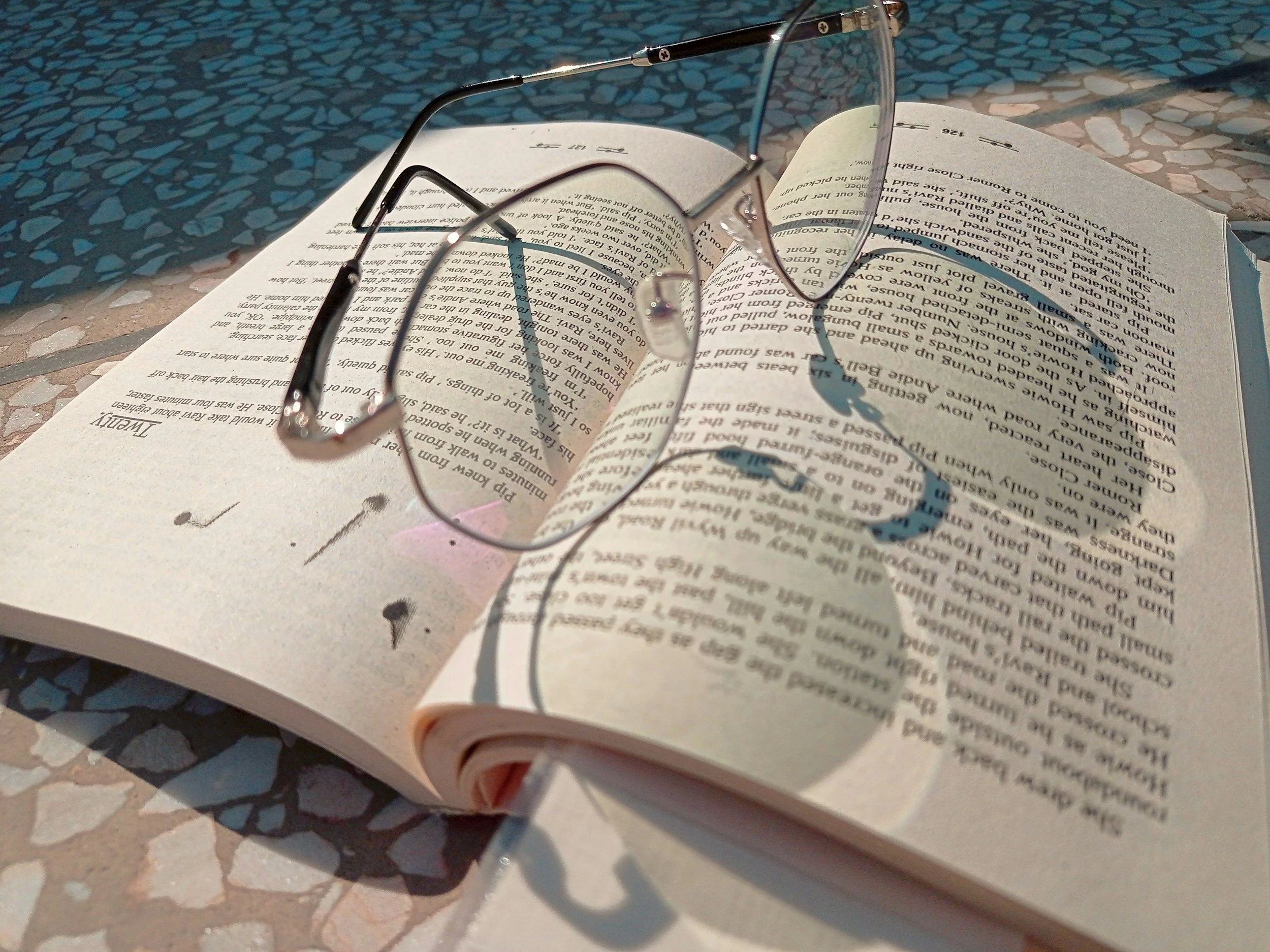 An open book with reading glasses placed on top, casting shadows on the pages, on a textured surface.