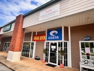 Exterior view of a shopping plaza with a Costa shop and an optical center, with signs indicating 'Now Open' and 'Open' on the storefronts.