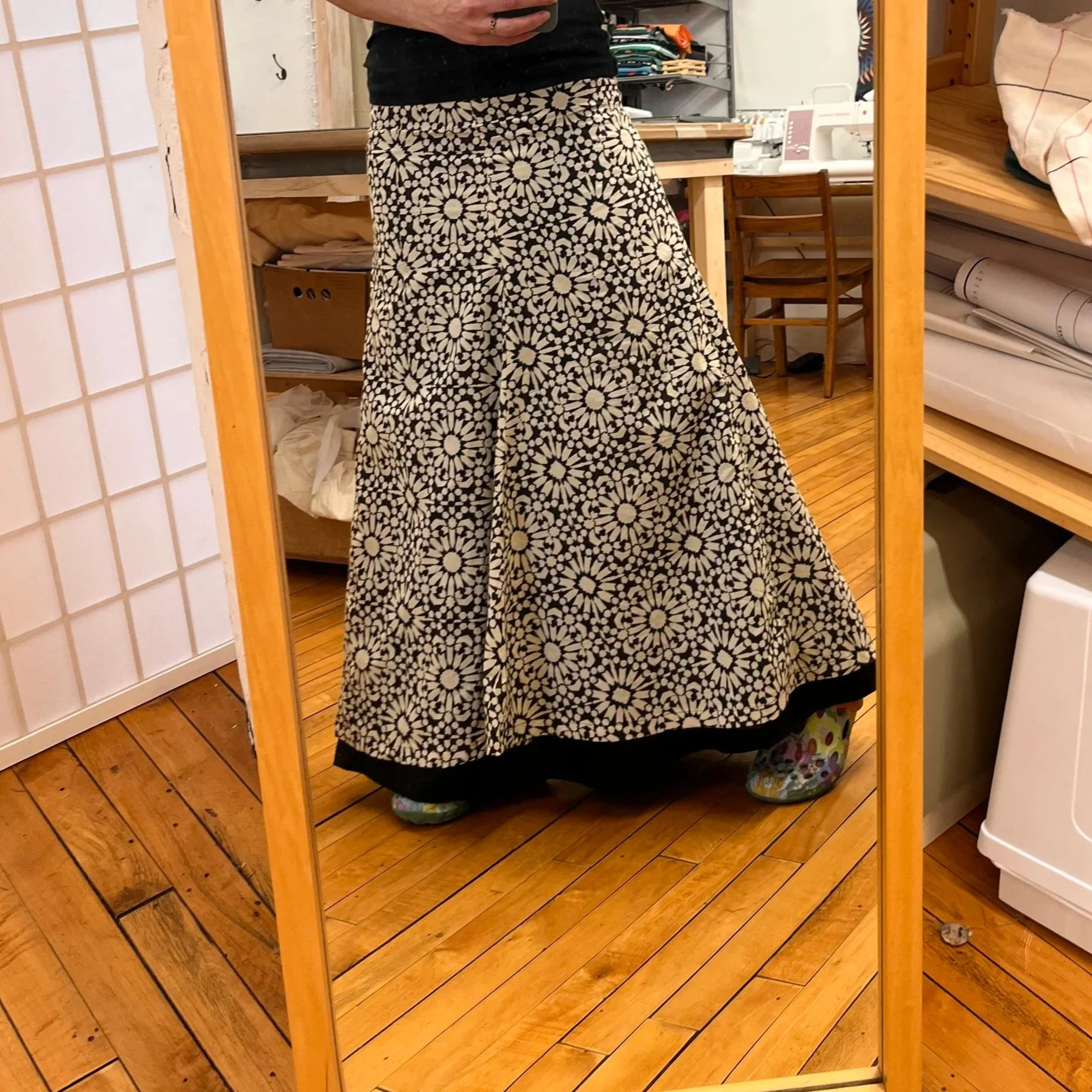 Person taking a mirror selfie, wearing a long black top, wide-legged floral skirt, and colorful shoes, standing on a wooden floor.