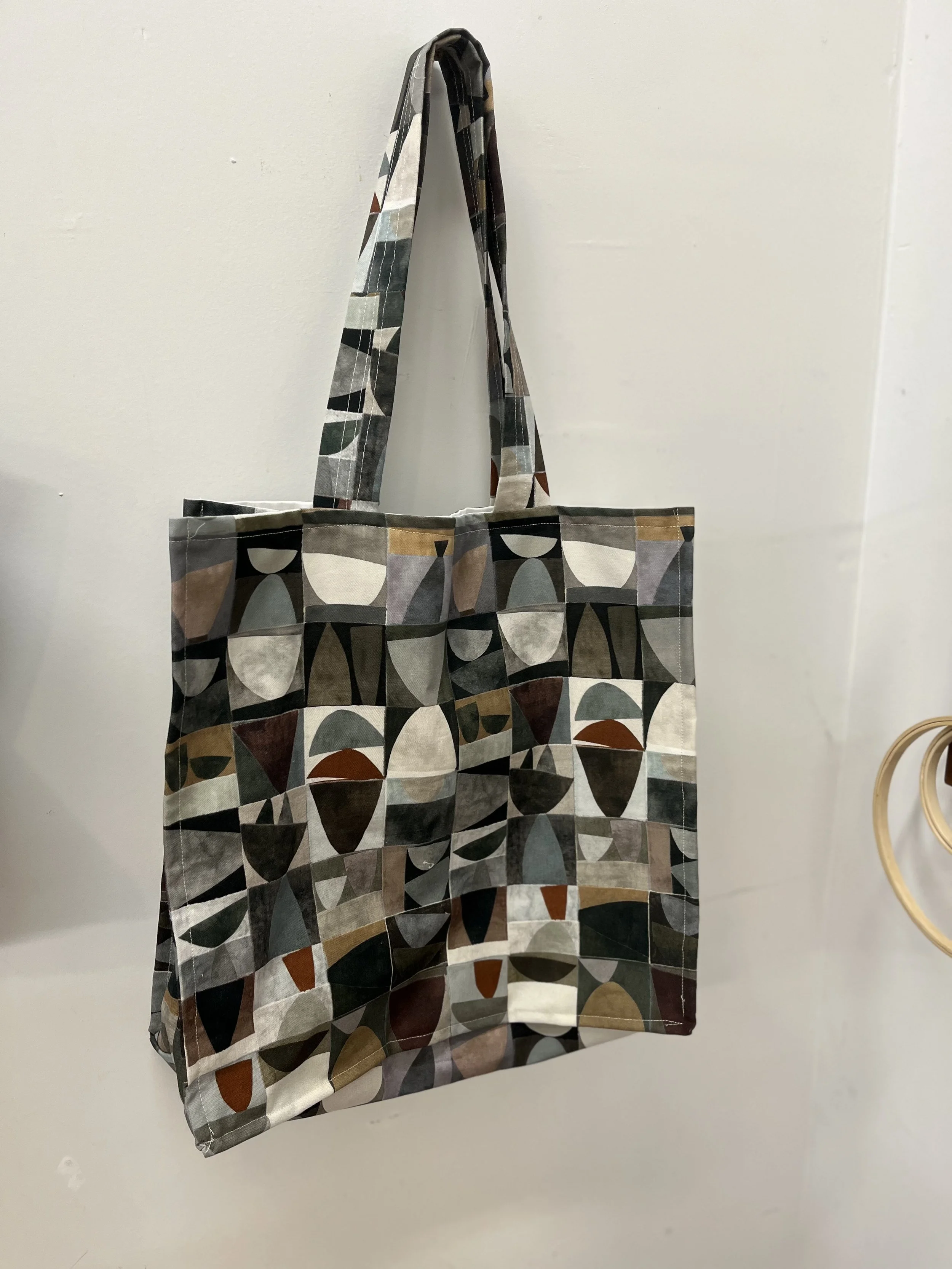 A fabric tote bag with a geometric pattern of various colors, hanging on a wall.