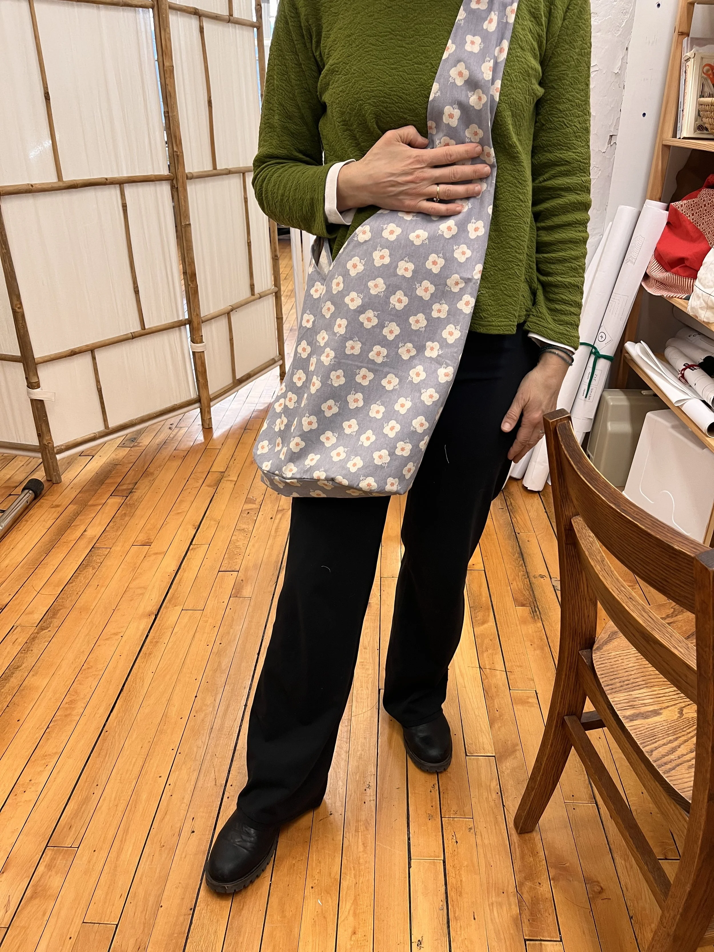 Person standing indoors holding a fabric bag with a floral pattern, wearing a green quilted jacket, black pants, and black shoes, with a wooden floor and a room divider in the background.