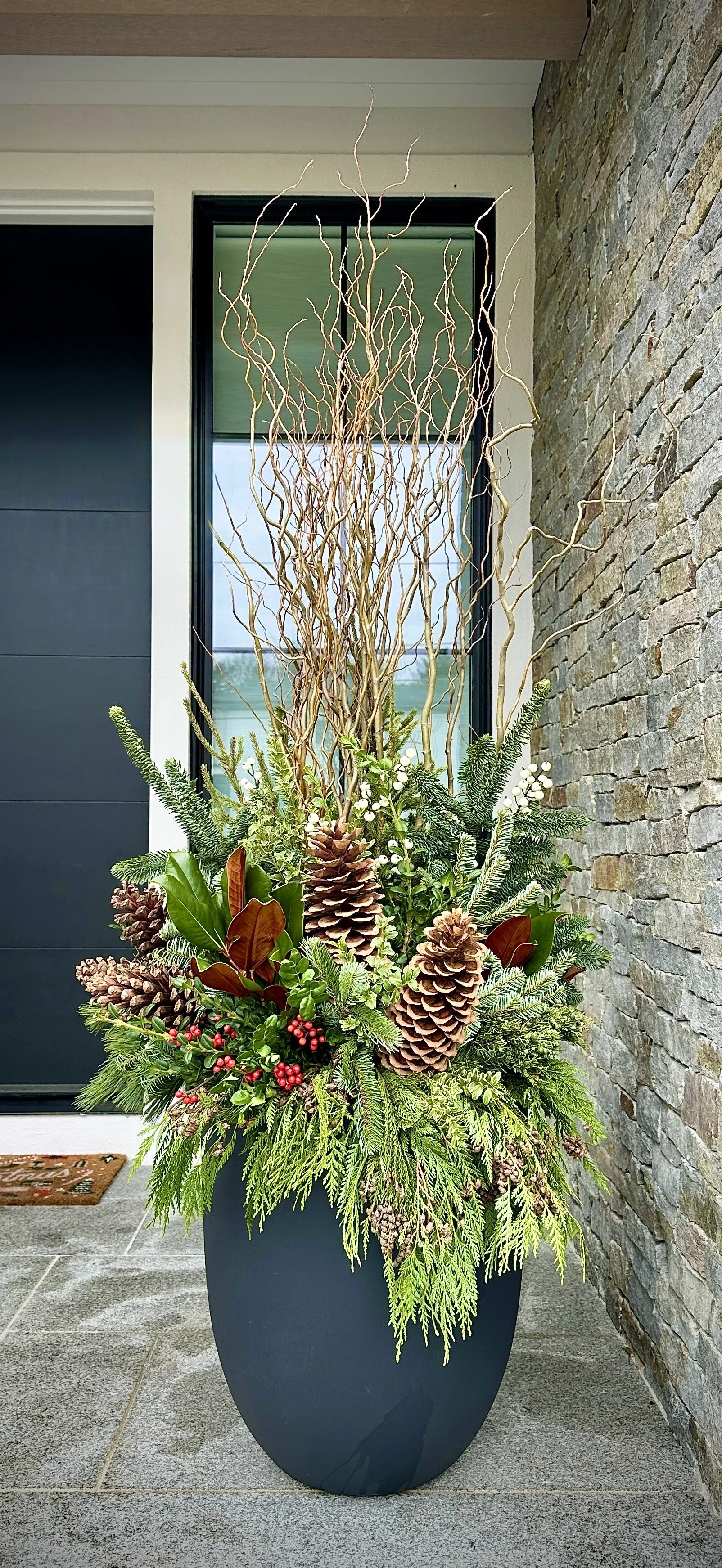 Decorative holiday plant arrangement, featuring pinecones, evergreen branches, white berries, and curly willow, placed at a front door.