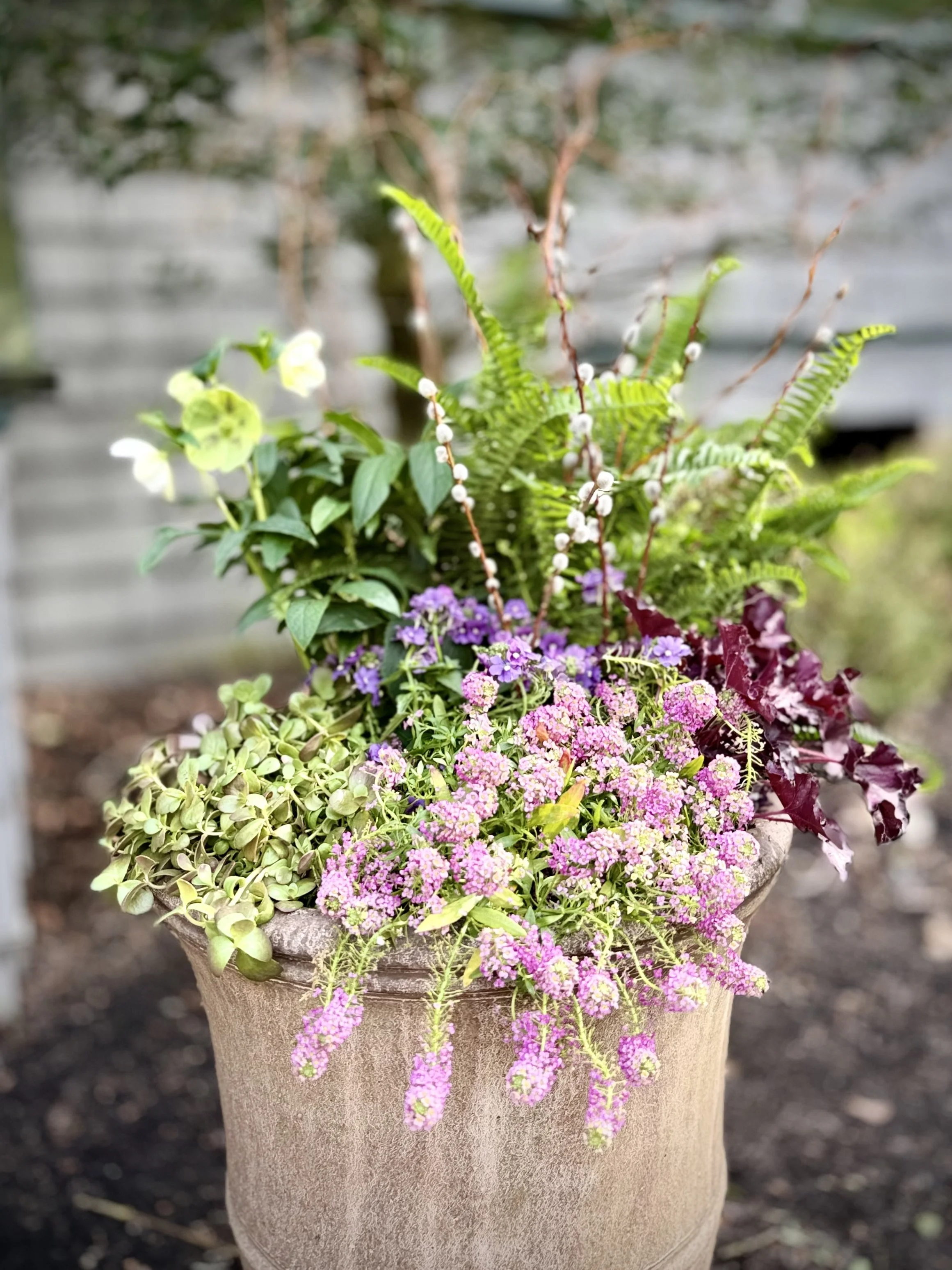 A large stone pot filled with assorted foliage and flowering plants, including pink and purple flowers, set outside an entryway.
