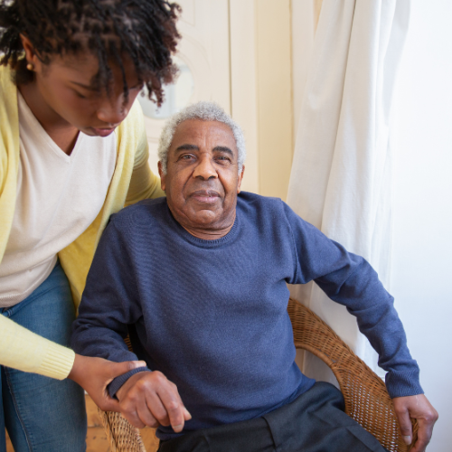 A young woman with natural curly hair assisting an elderly man with gray hair, sitting in a wicker chair near a window with white curtains.